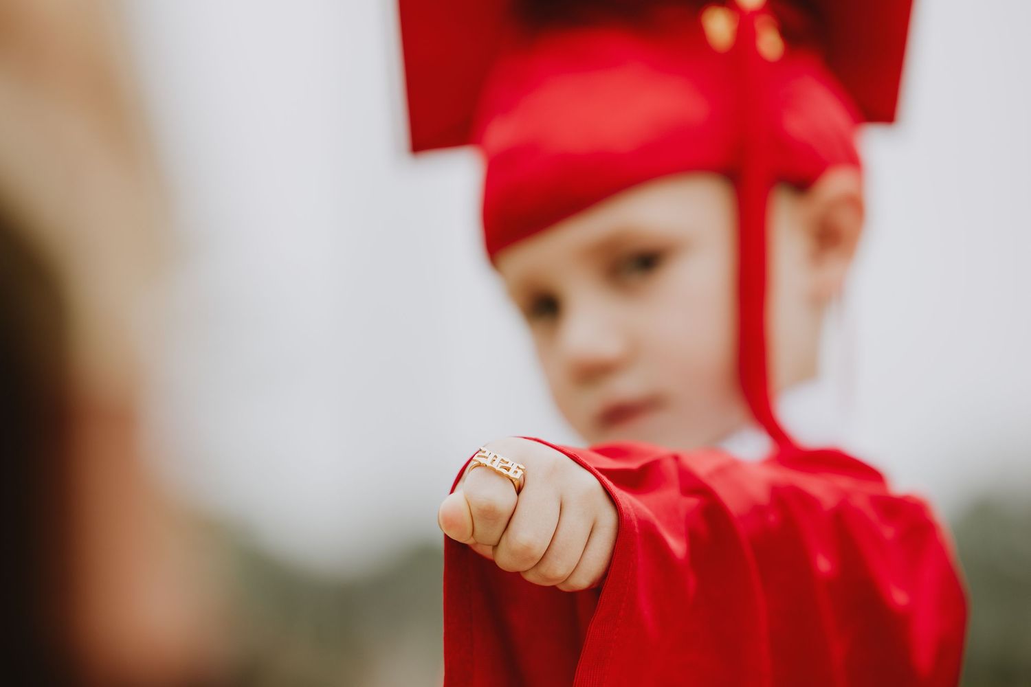 Young child in red graduation cap and gown pointing finger, preschool graduation ceremony outdoors.