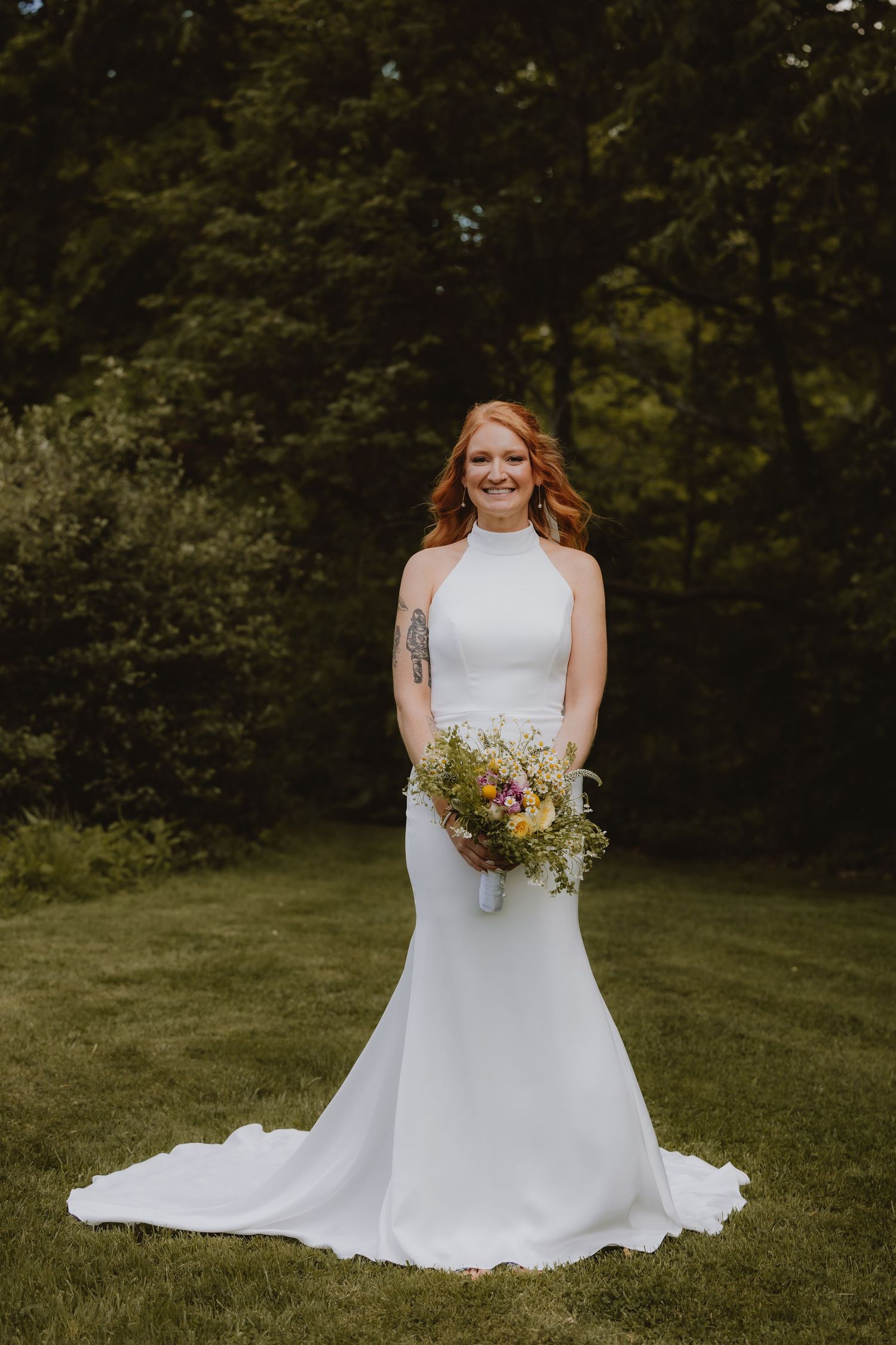 Bride in white halter mermaid gown holding colorful wildflower bouquet, standing on green lawn with lush trees behind.