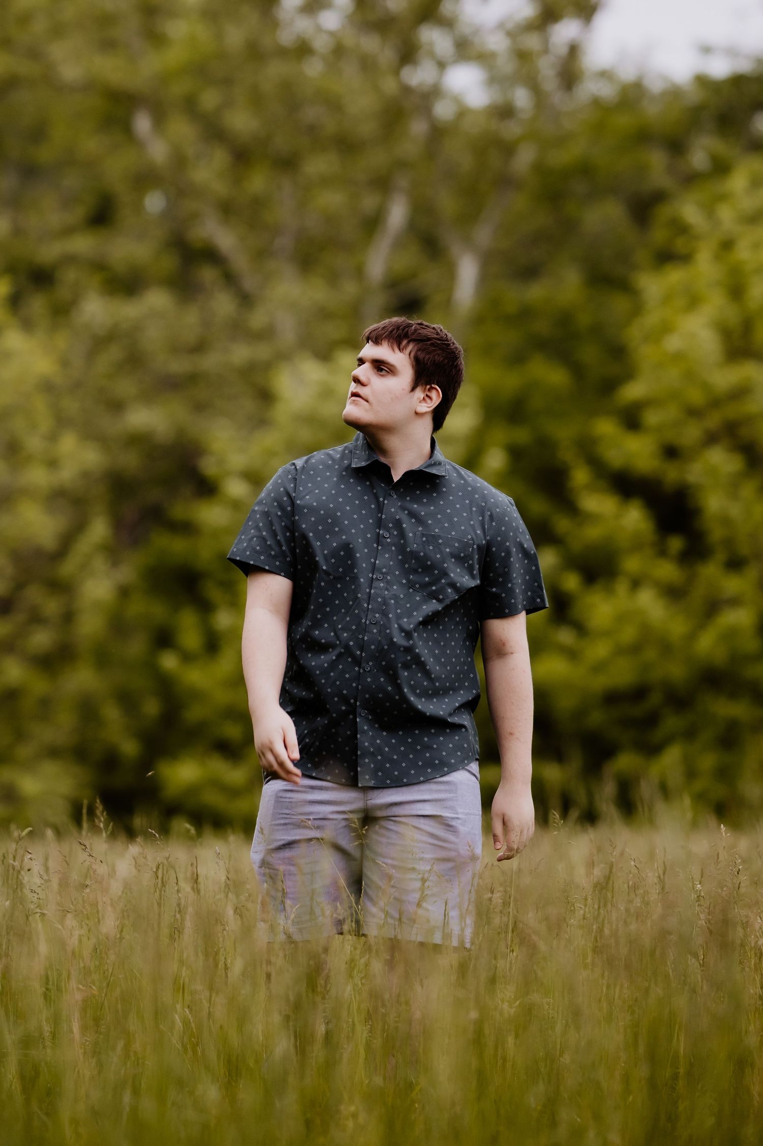 Young man in dark patterned shirt and khaki shorts standing in tall green grass field with trees in background.