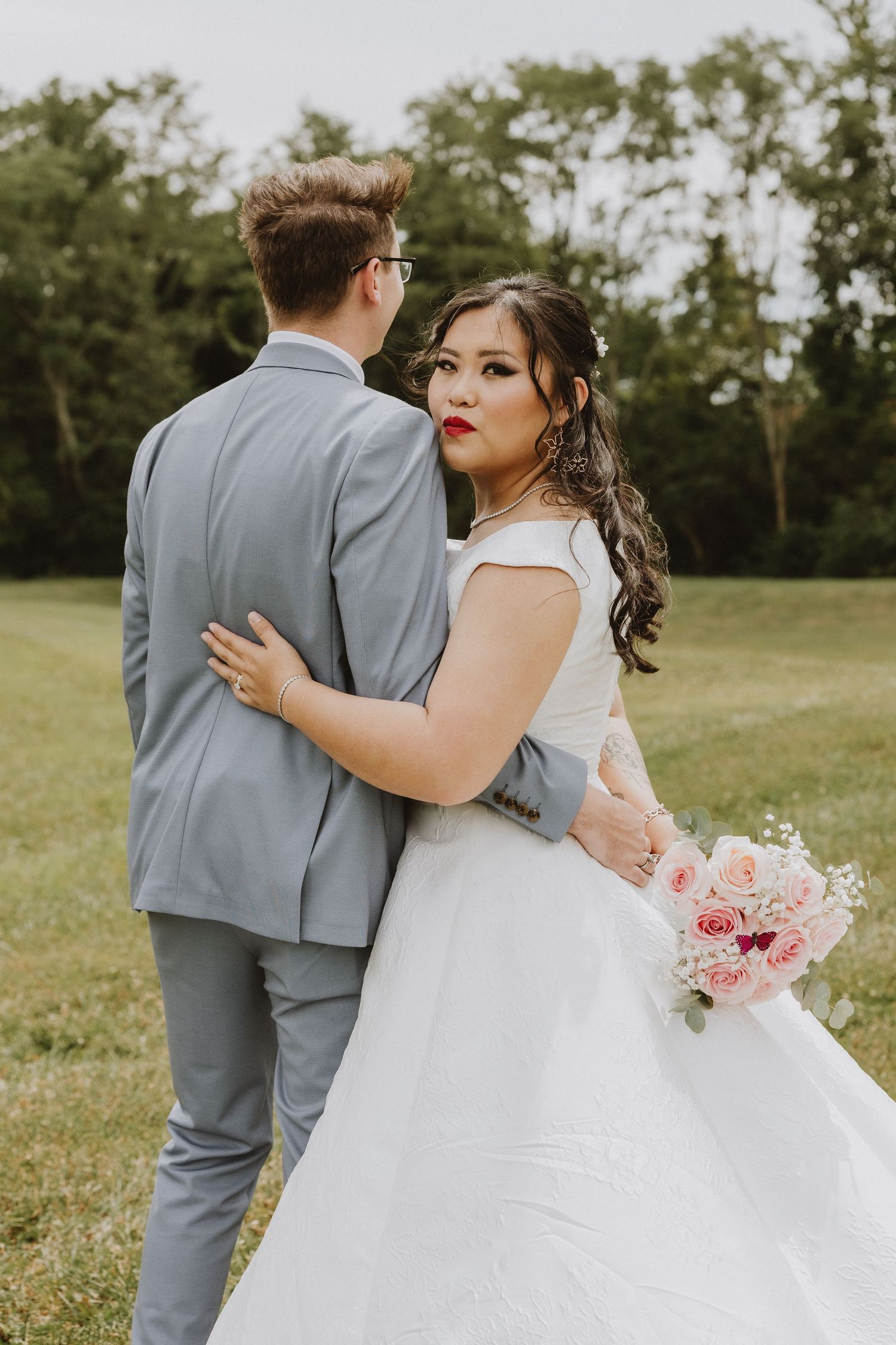 An intimate wedding portrait with soft pink bouquet and classic grey suit against green landscape.