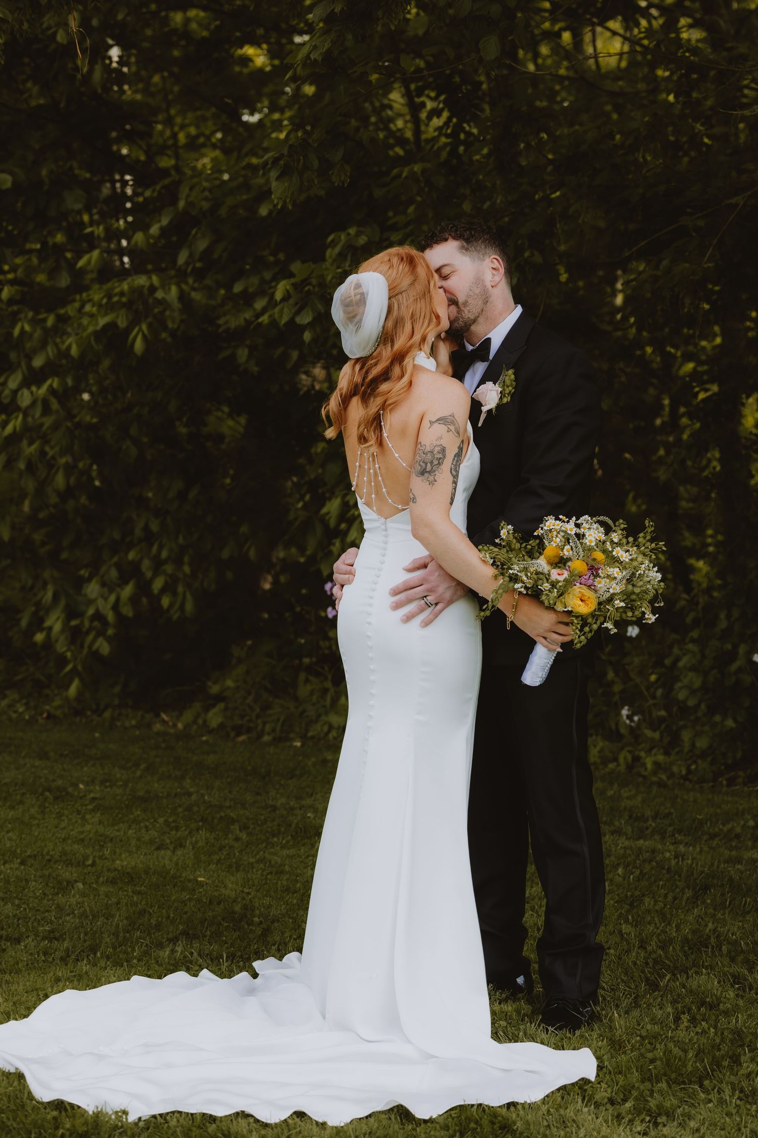 Bride and groom share a kiss outdoors, she wears a white gown with train, he wears a black tuxedo, holding wildflower bouquet.