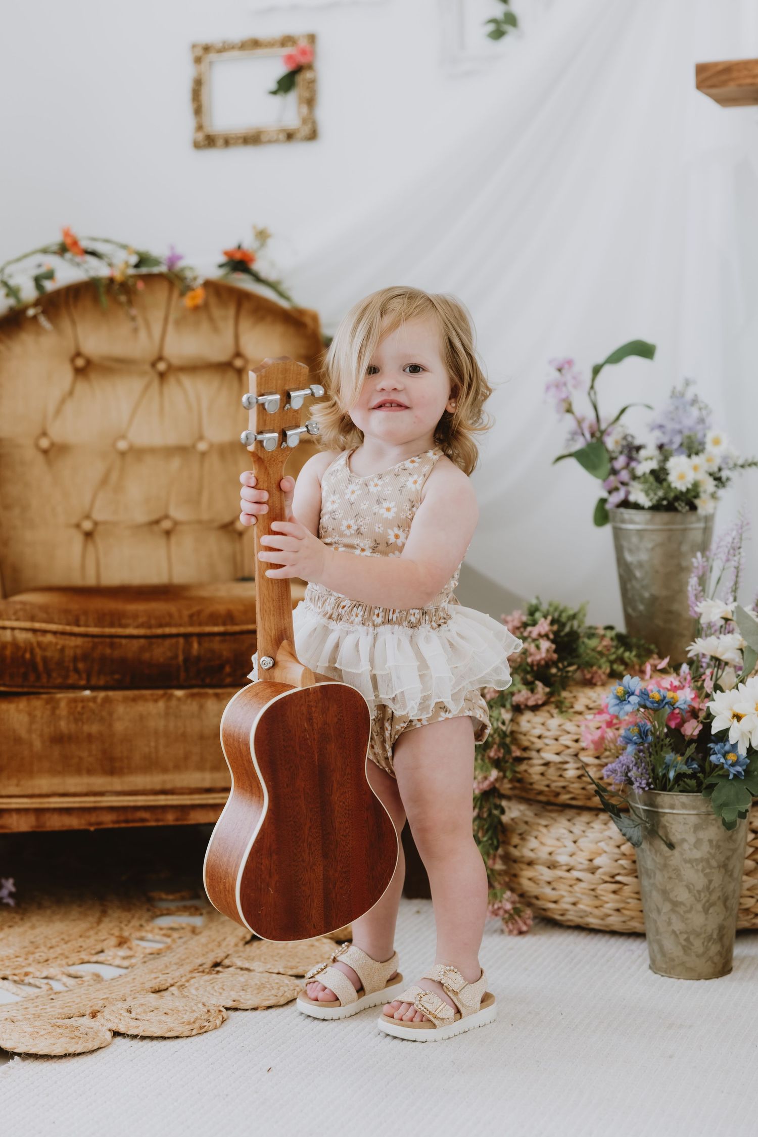 Toddler girl in floral romper holding acoustic guitar beside vintage sofa with flower arrangements.
