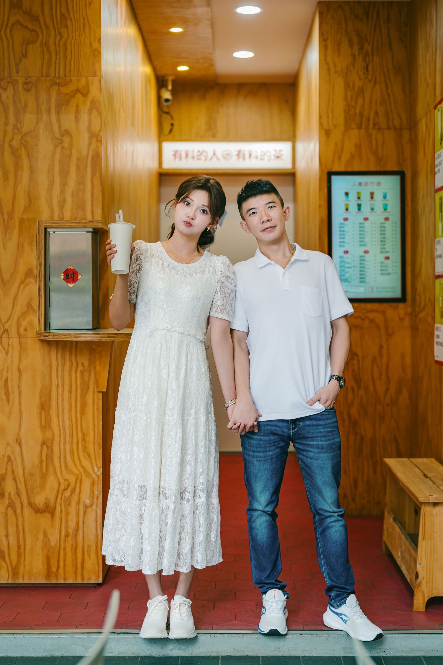 A couple stands in a hallway with wooden paneled walls, wearing casual white outfits and sneakers.
