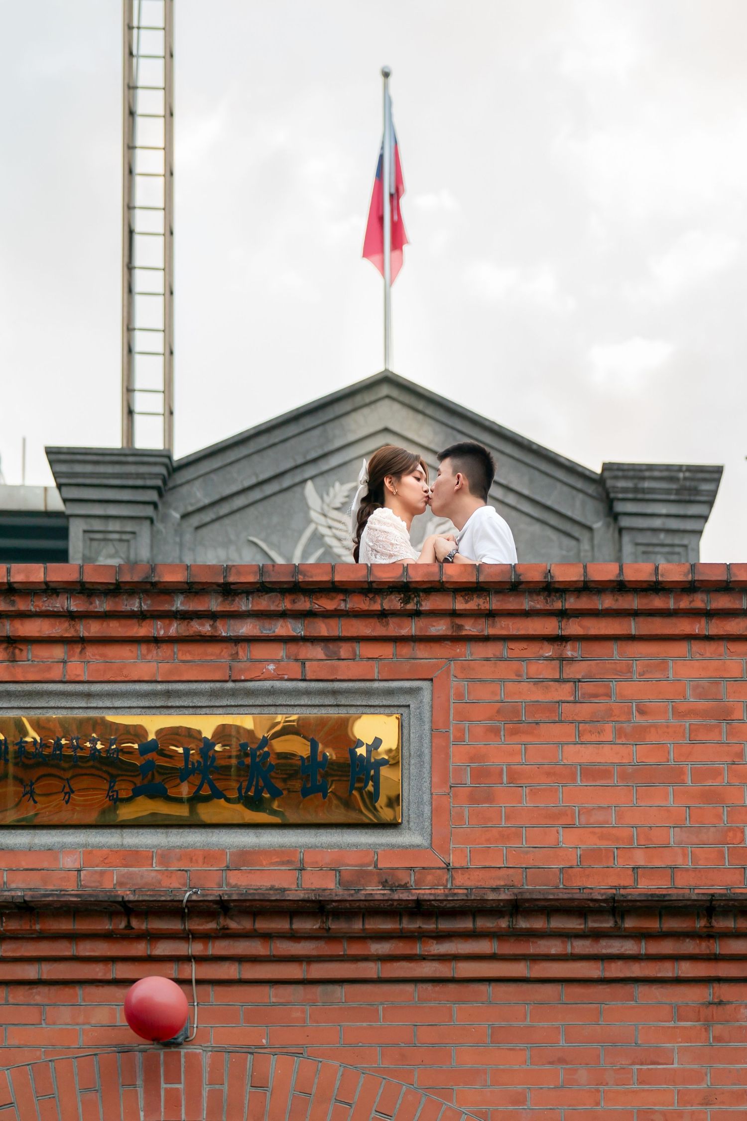 Red brick wall with green gabled roofline and flags mounted on top under cloudy sky.