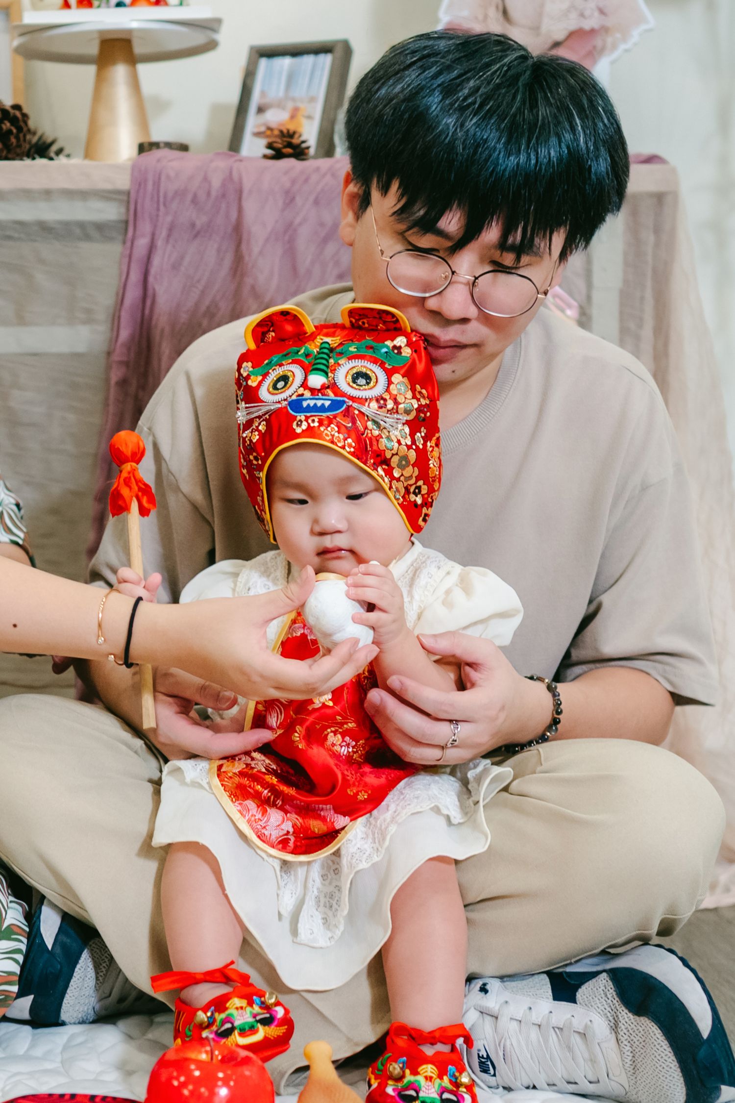 Baby wearing traditional red Chinese New Year outfit with matching hat and booties sits with caregiver in casual grey attire.