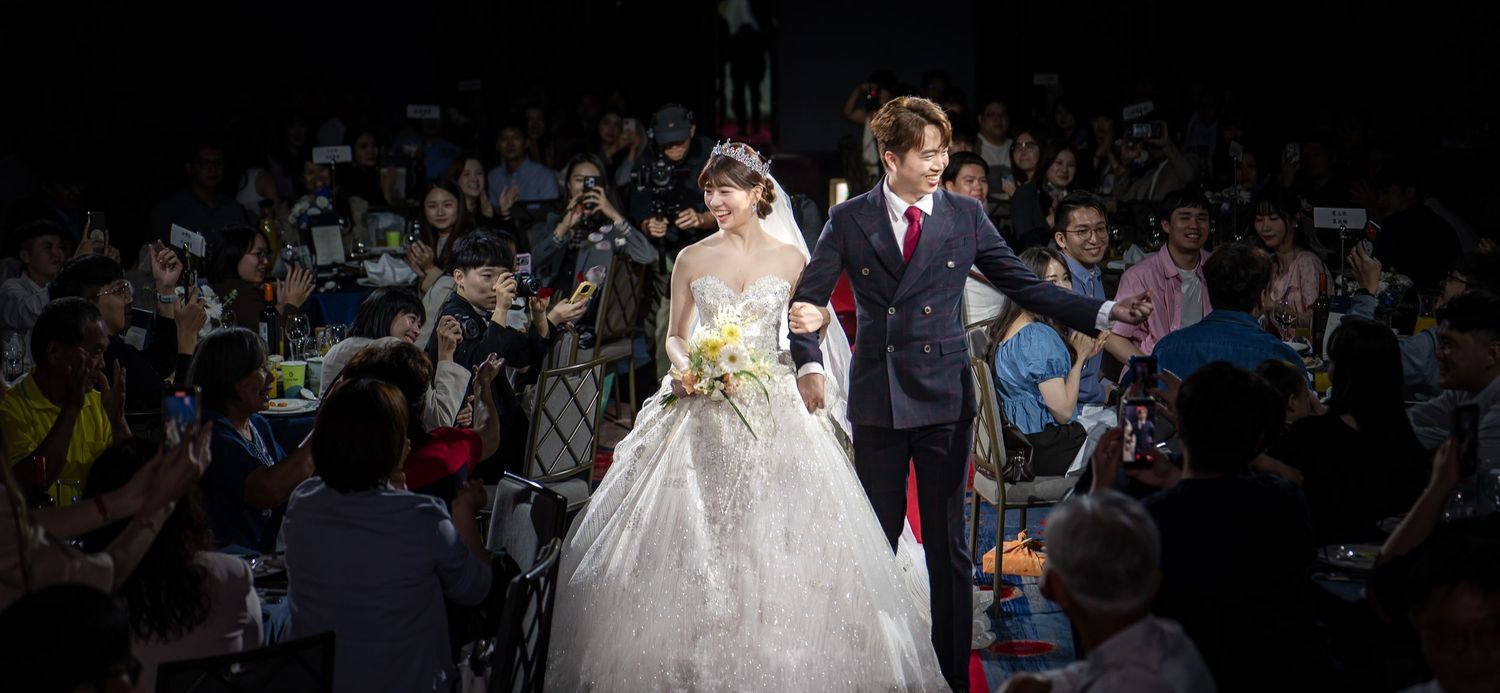 Bride in white ball gown and groom in navy suit walk down aisle at elegant wedding ceremony with guests.