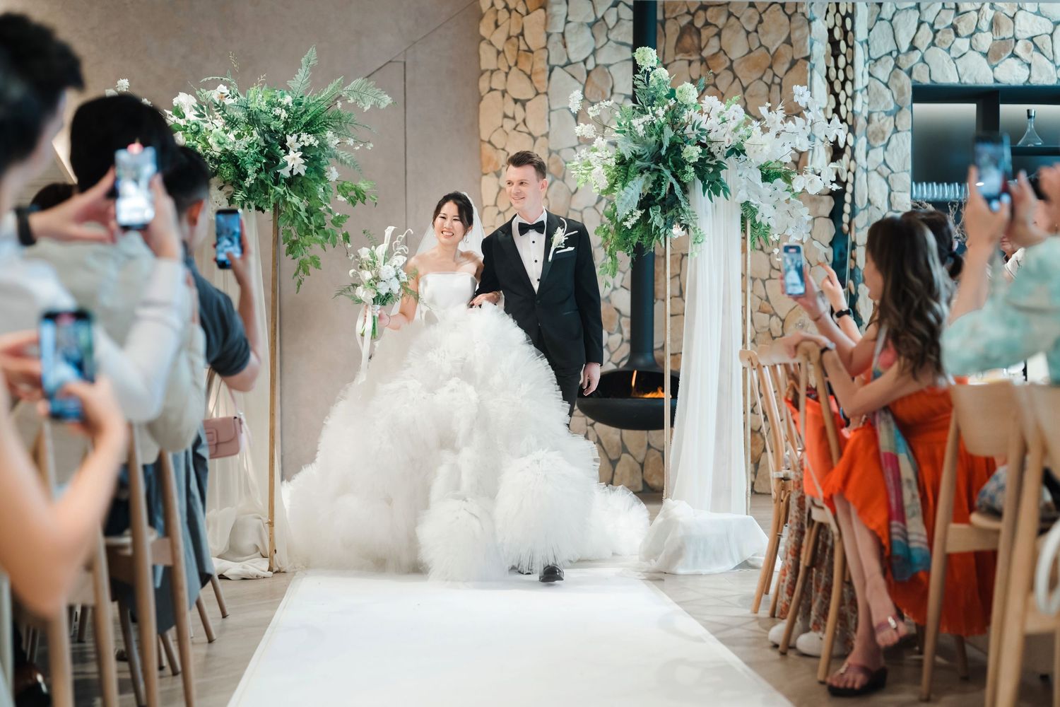 Wedding ceremony in progress with photographers capturing the moment as couple walks down aisle with white floral decorations.