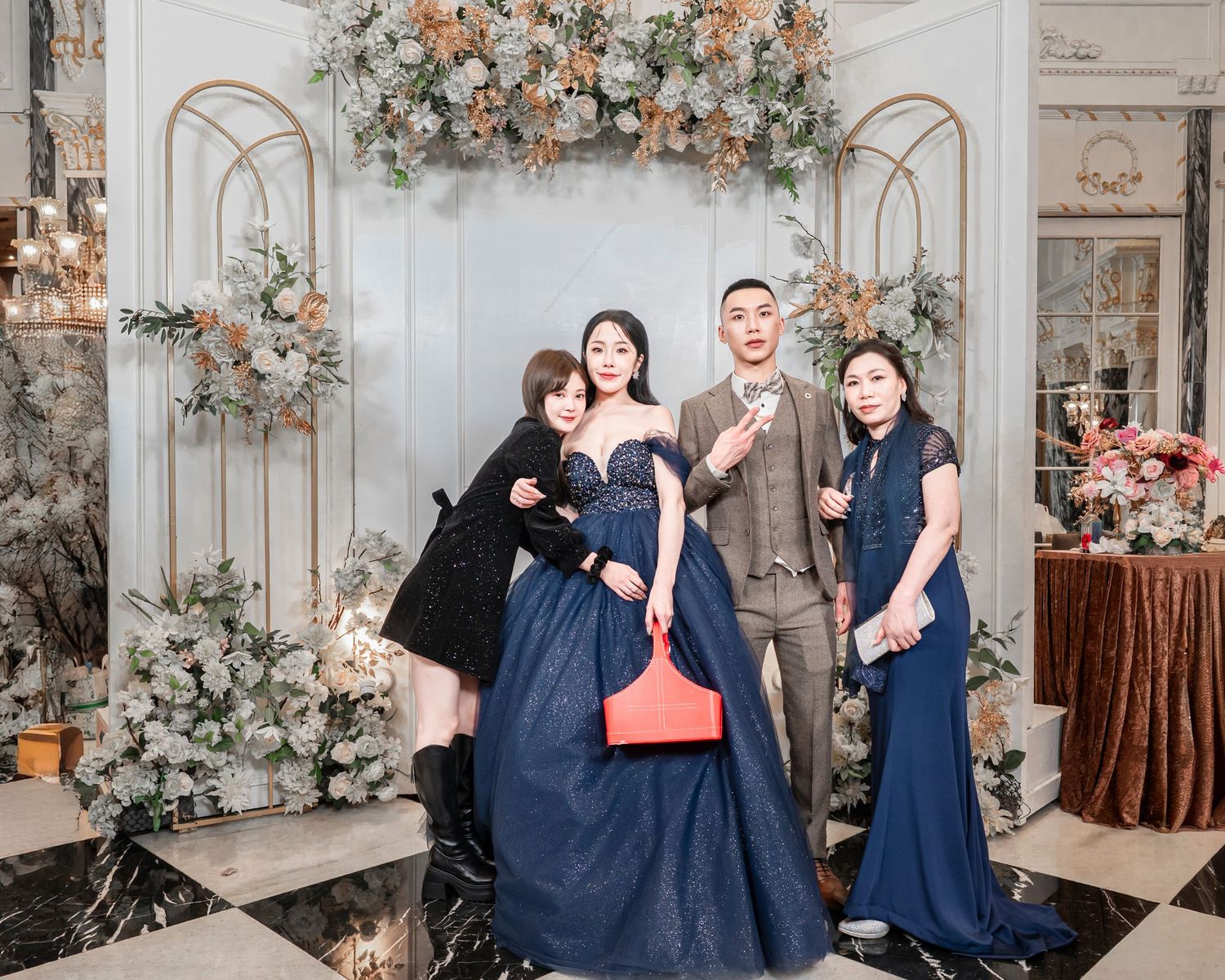 A wedding party in navy blue dresses and suits poses in front of an elegant floral arch with peach and white flowers.