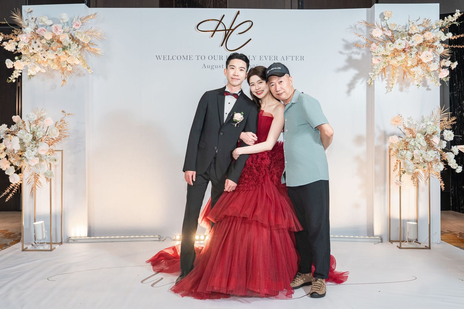 Wedding celebration scene featuring formal attire with elegant red ball gown against white backdrop with floral decor.