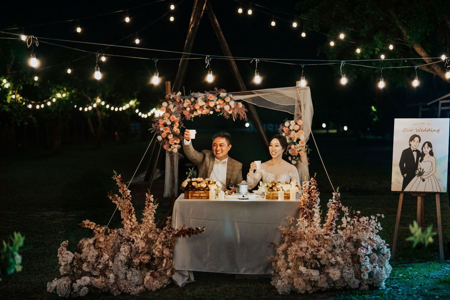 Outdoor wedding reception setup with twinkling string lights and pink floral decorations at dusk.