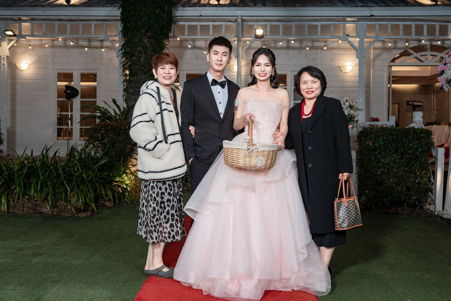 Wedding party poses together in front of an elegant venue with glass windows and greenery at dusk.