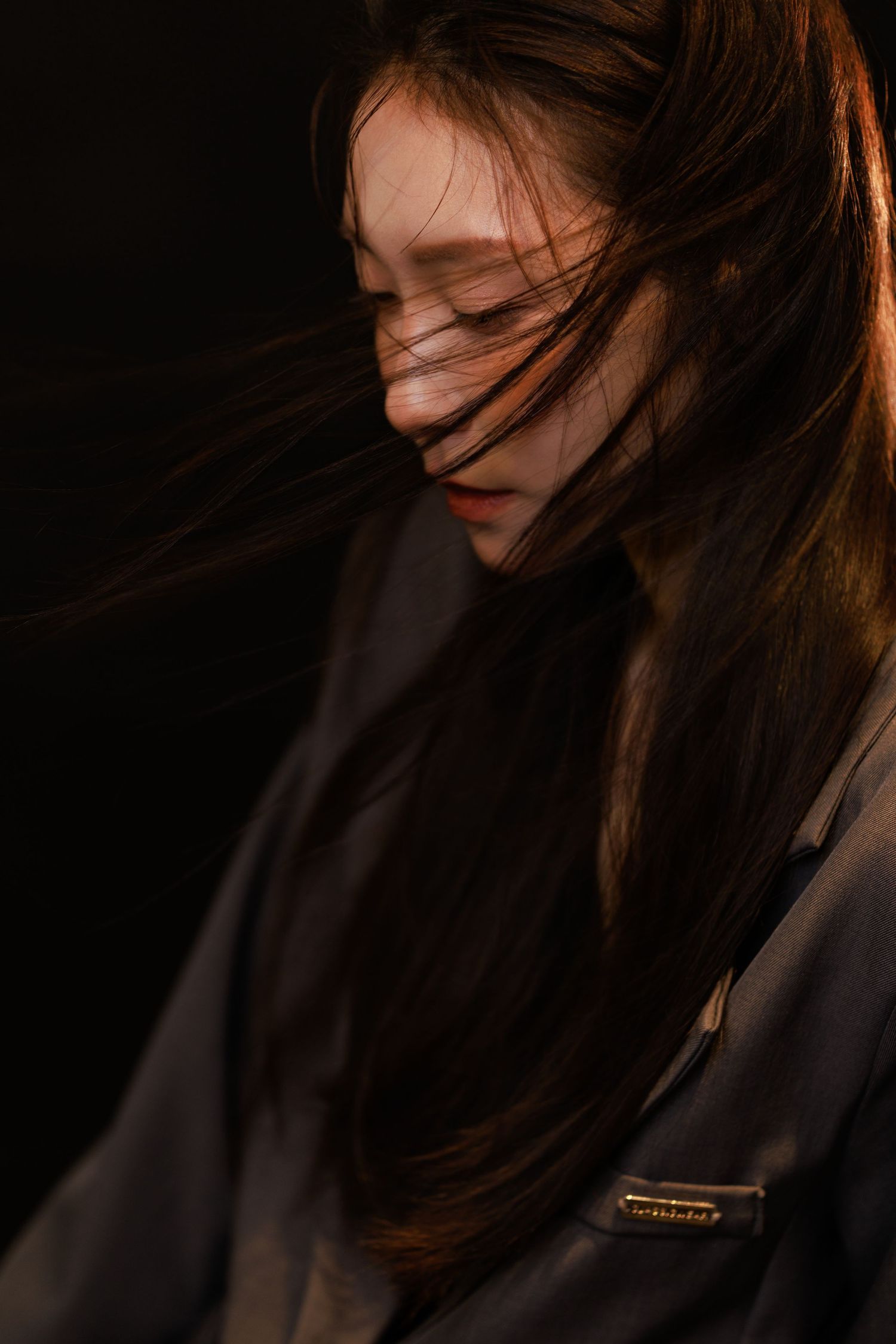 Young woman with long dark hair blowing across her face, eyes closed, dramatic dark background with warm lighting.