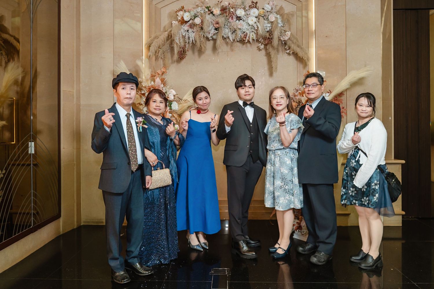 A group of people dressed in formal attire pose together under an elegant floral arch at a special event.