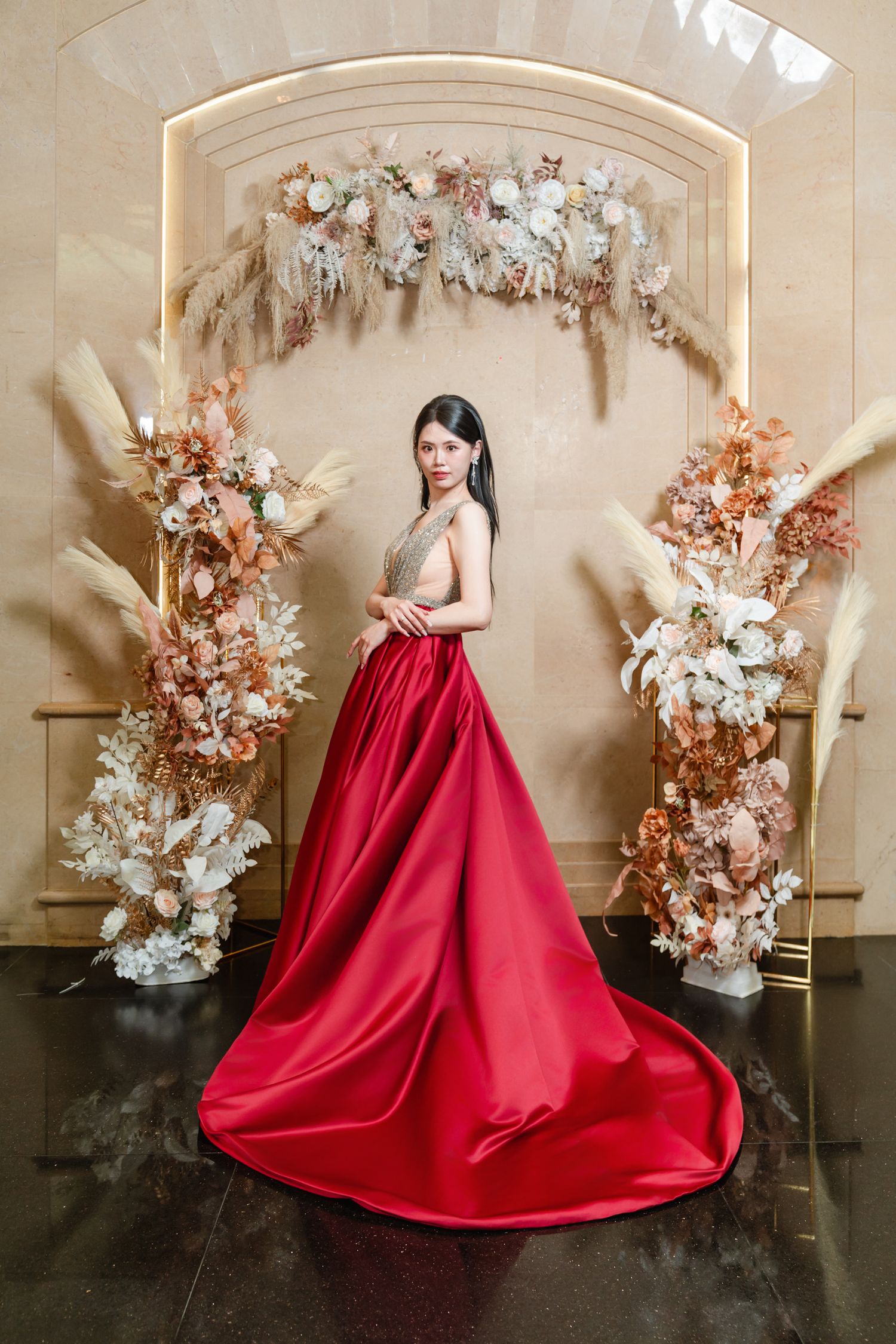 Elegant red ballgown in front of floral arch with dried white and copper flowers against beige wall.
