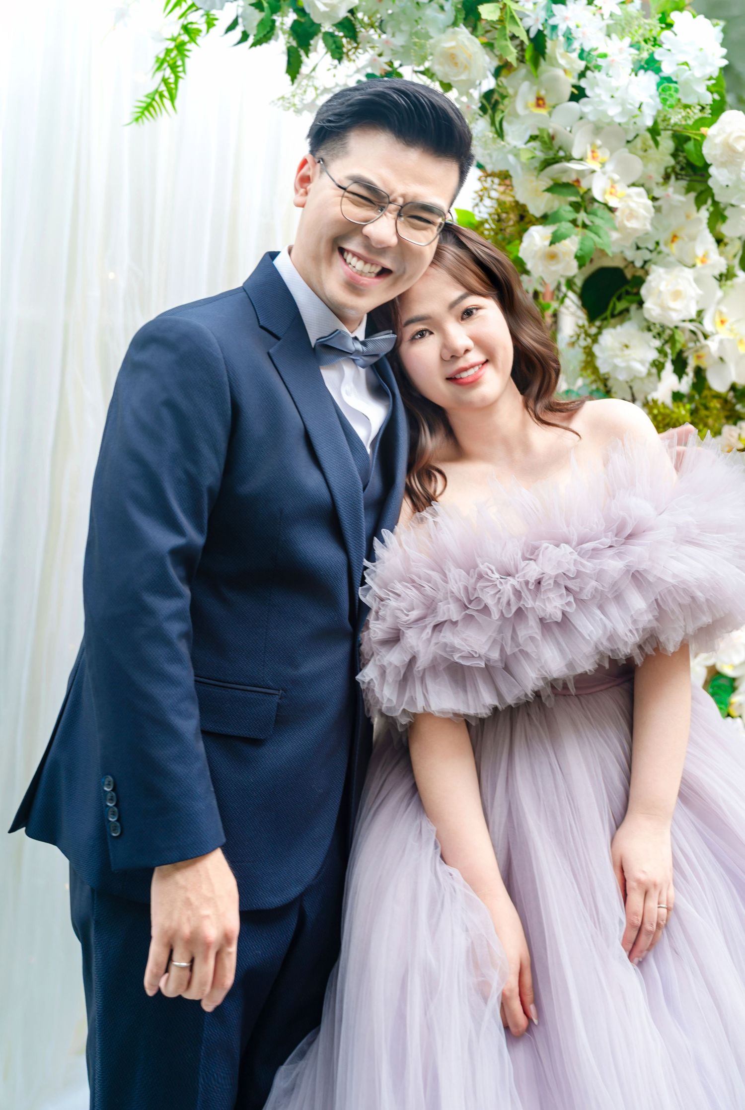 A couple poses together in formal attire, with a navy suit and soft lavender tulle gown against a floral backdrop.
