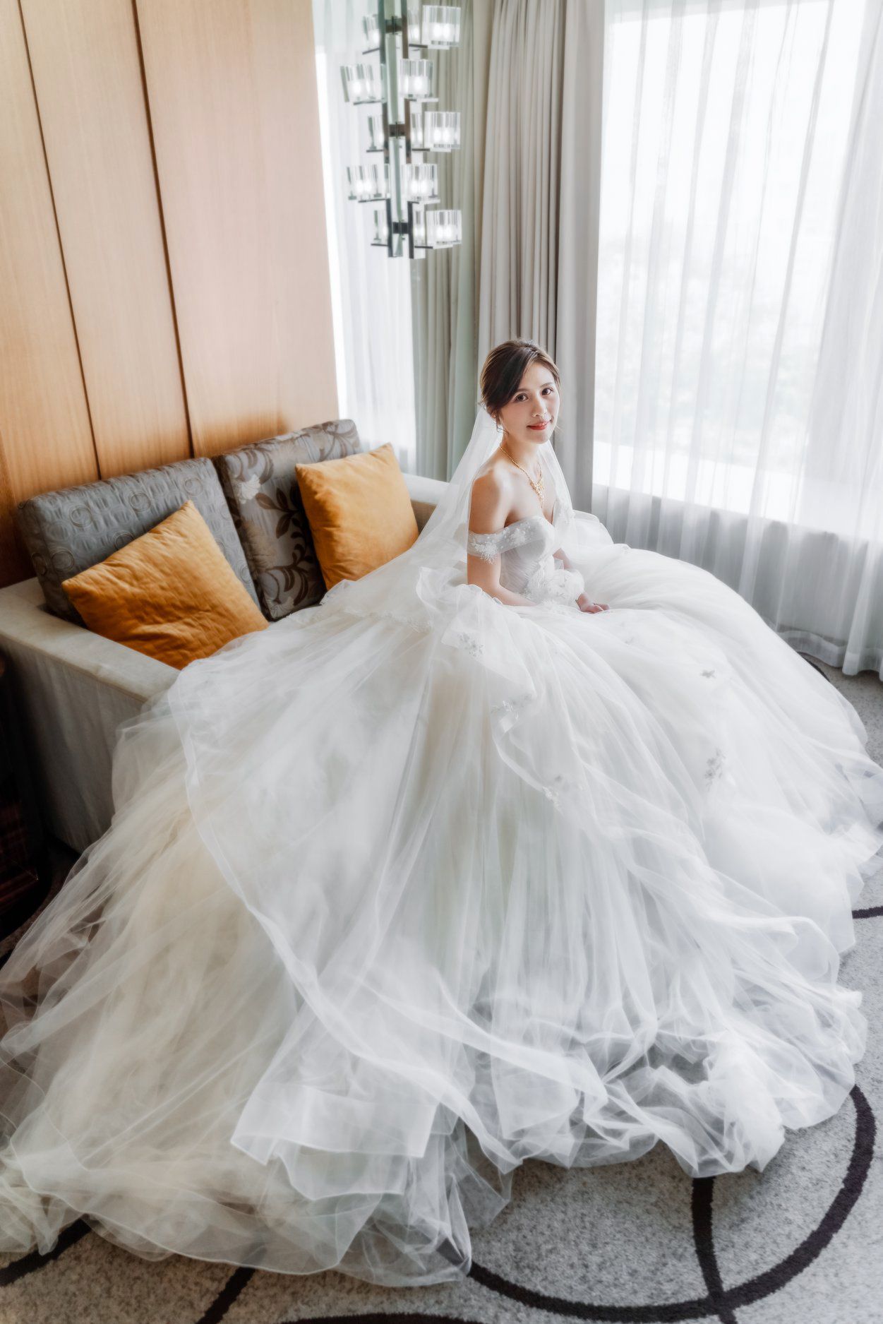 A bride in an elegant white tulle ballgown poses gracefully on a gray sofa with golden accent pillows near sheer curtains.