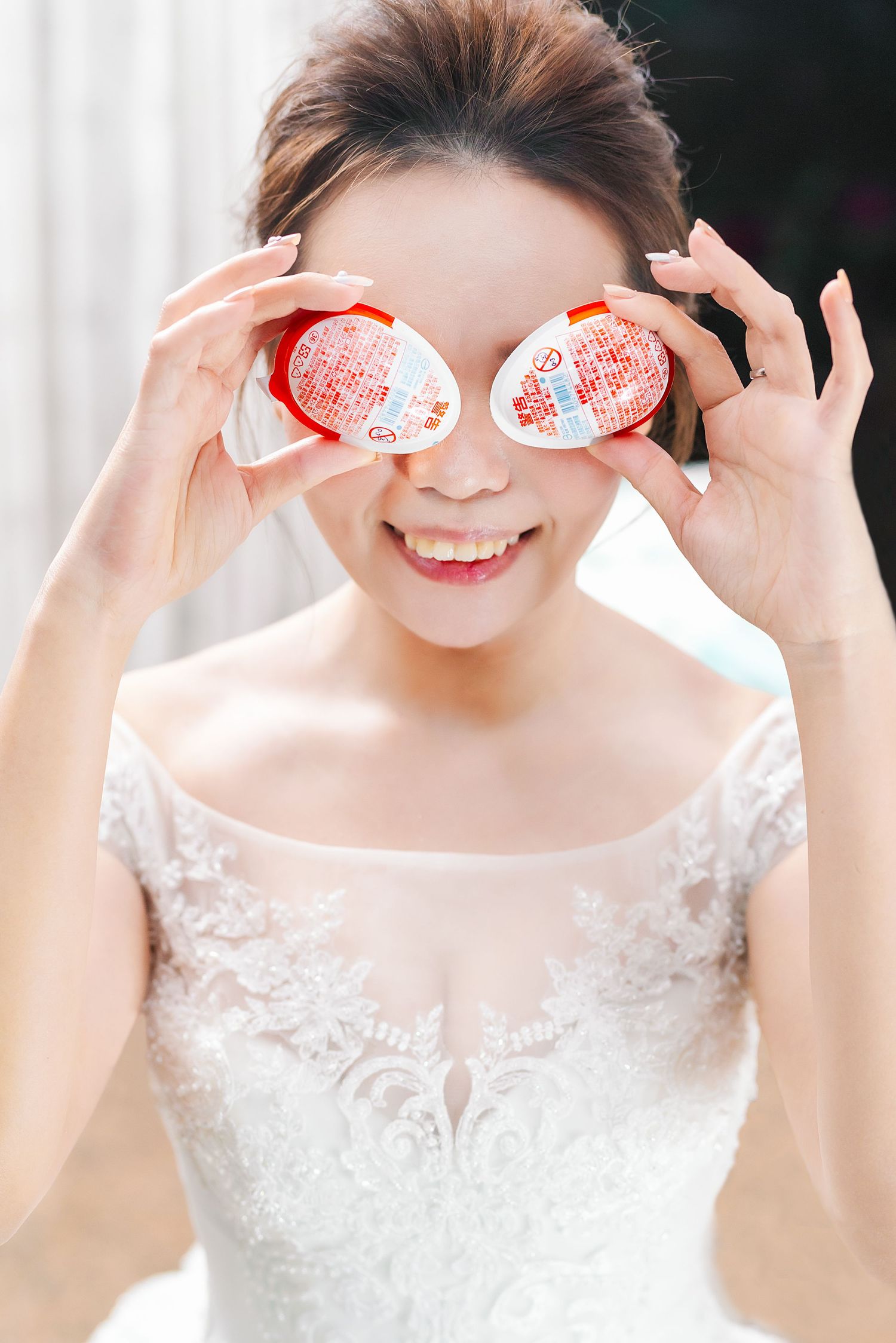 Person in white lace wedding dress holds traditional red double happiness symbols over eyes in playful pose.