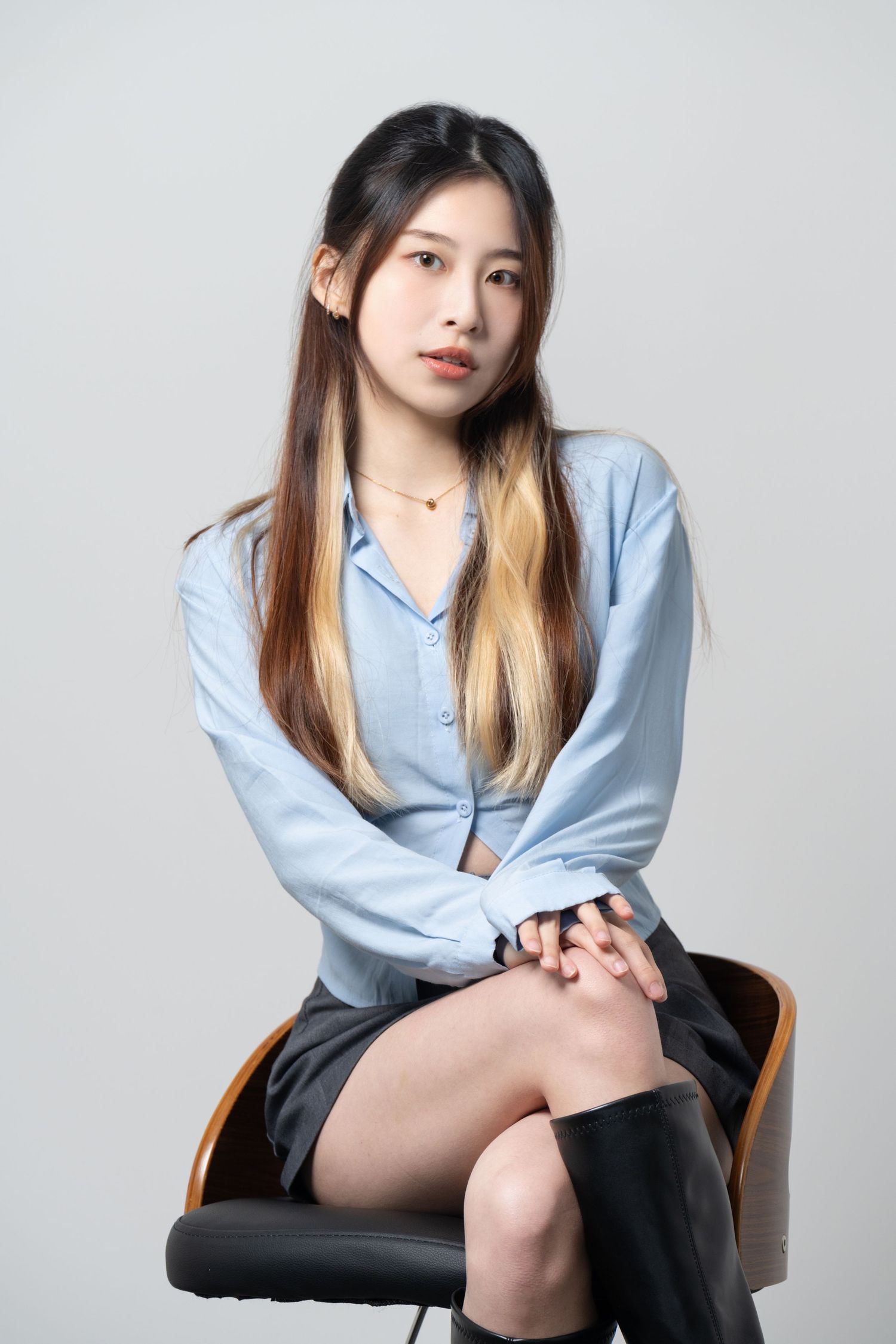 Portrait of someone wearing a light blue button-down shirt and black skirt seated on an office chair against gray background.