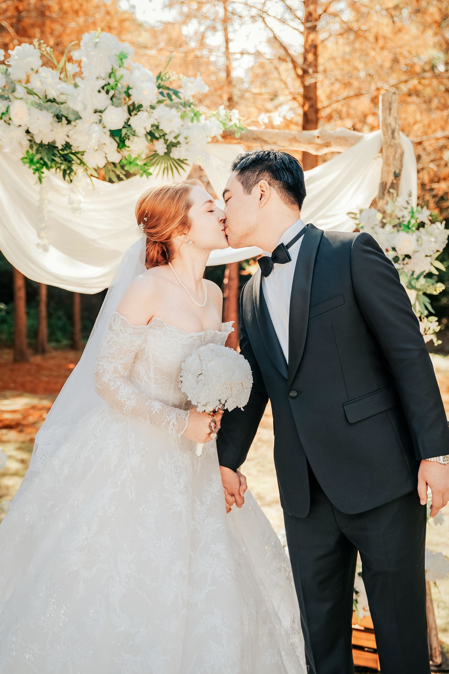 A romantic autumn wedding scene with a white floral arch, flowing drapery, and a couple sharing a kiss in formal attire.
