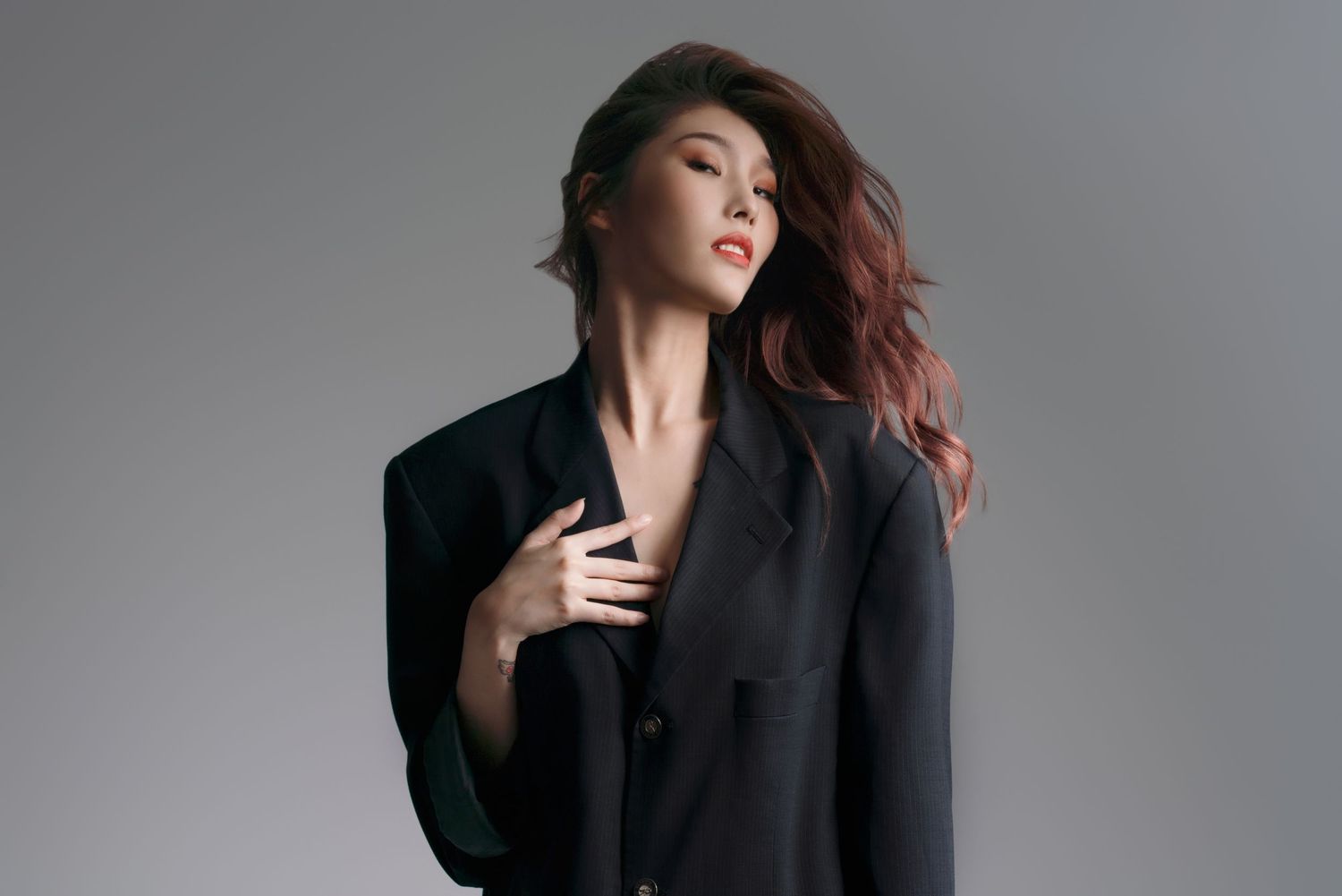 Model in black blazer posing dramatically against white background with wind-blown red hair.