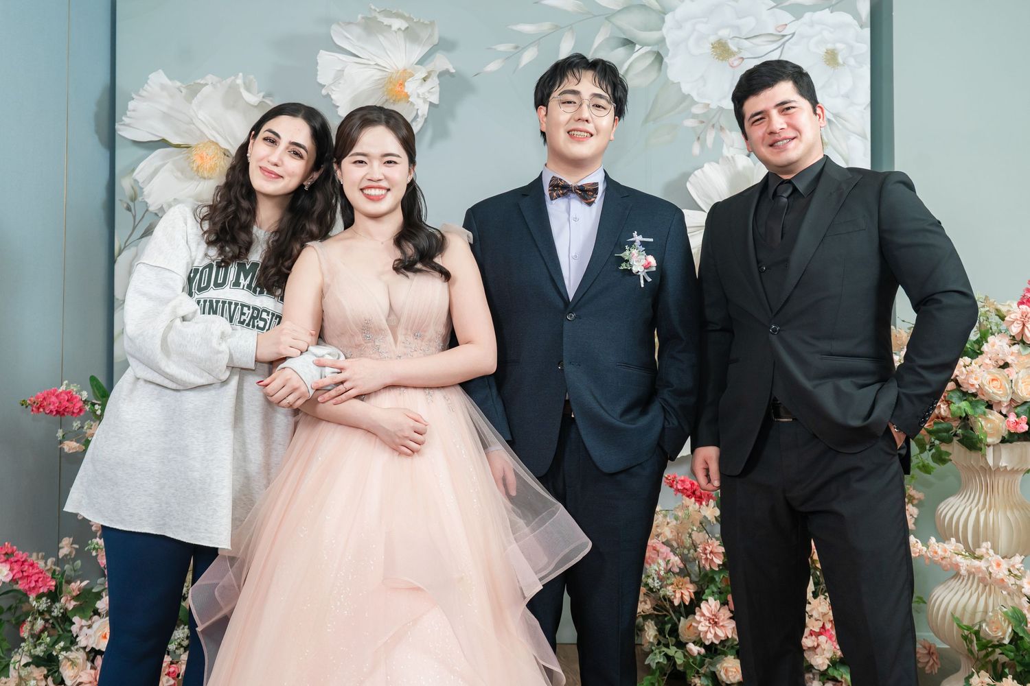 Wedding party members pose together in formal attire with floral decorations in the background.