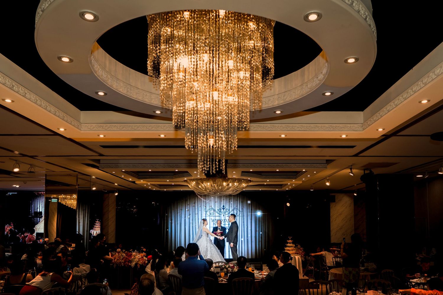 A grand ballroom wedding ceremony under a crystal chandelier with guests seated at elegant tables watching the stage.