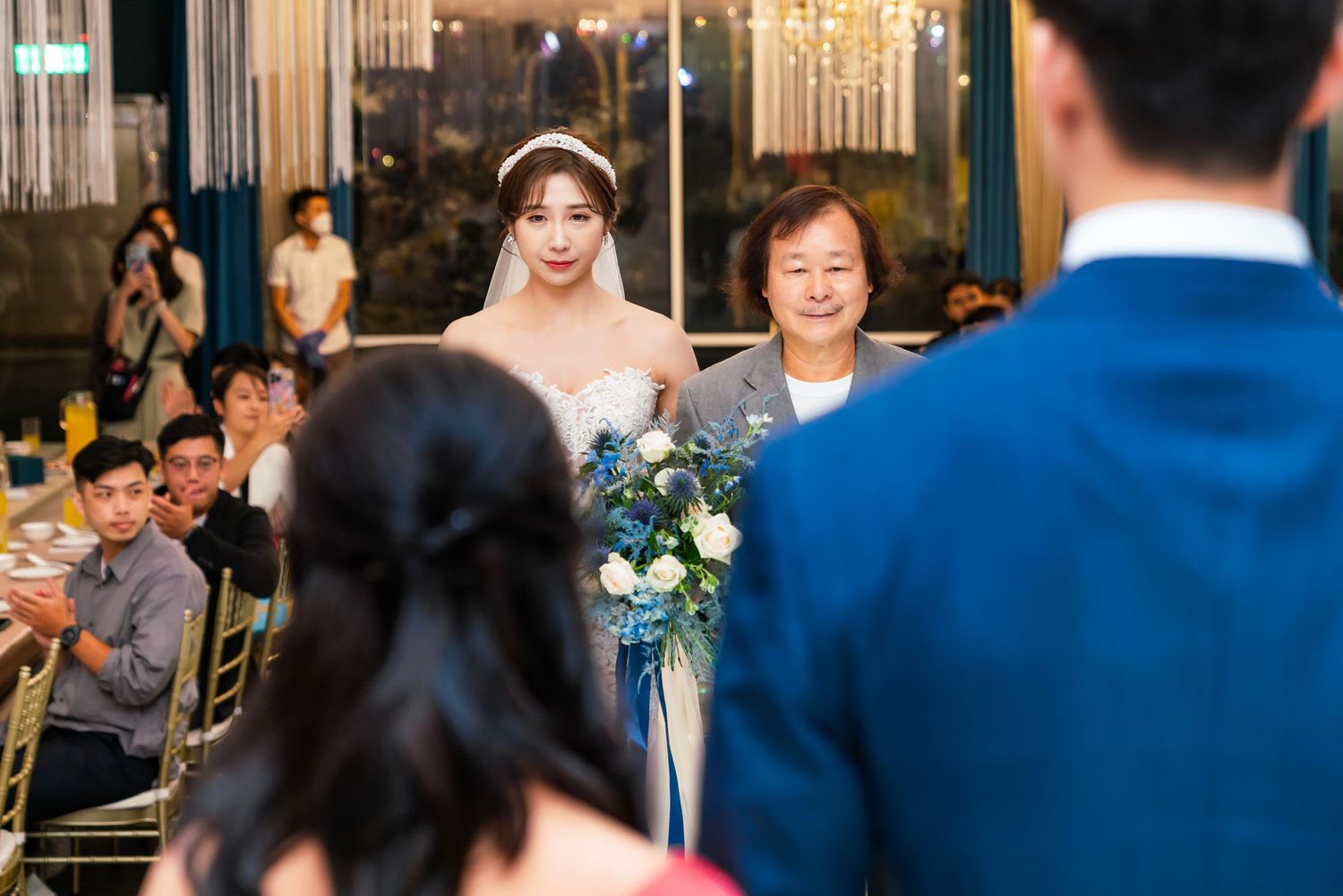 A bride and family member stand together at an elegant indoor wedding ceremony with chandeliers overhead.
