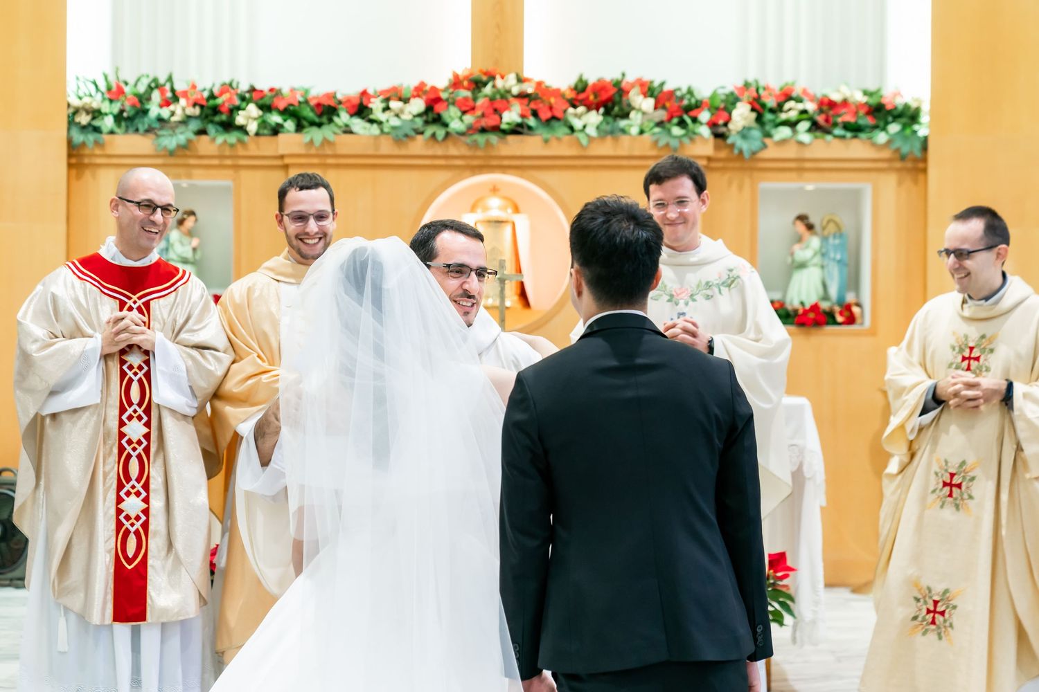 Priests in white robes officiate a wedding ceremony at a church altar decorated with floral garlands.