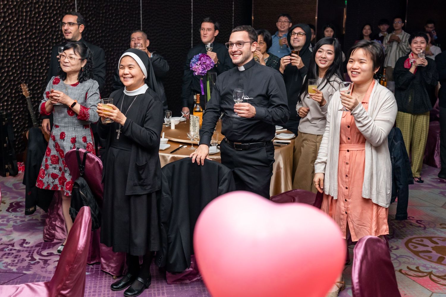 Wedding guests raise glasses during a toast with a pink heart balloon visible.