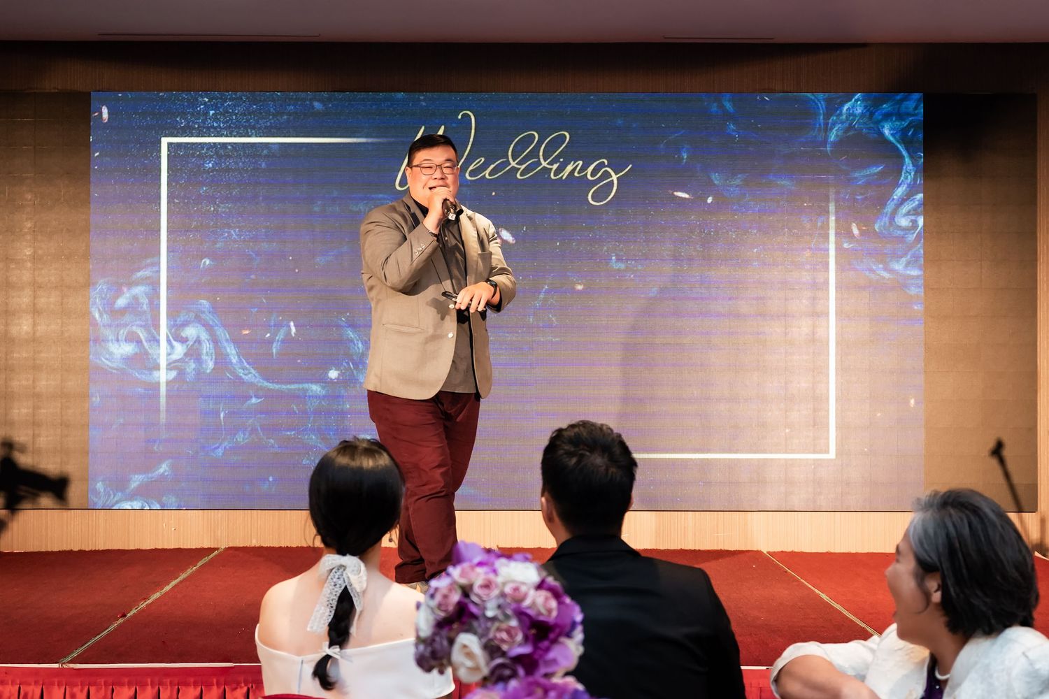 Presenter addresses seated wedding guests with purple florals and blue backdrop visible.