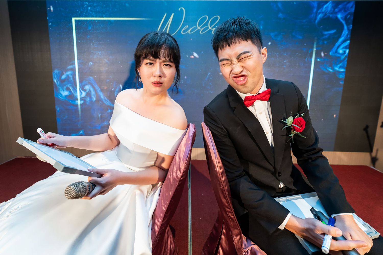 A couple in wedding attire sits on red chairs holding notebooks during their reception.