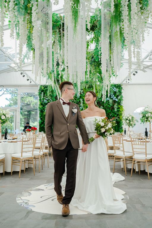 Couple stands in reception venue decorated with hanging greenery and white drapes.