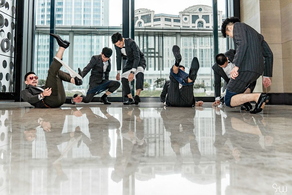 Wedding party members in suits pose playfully on a shiny marble floor near large windows.