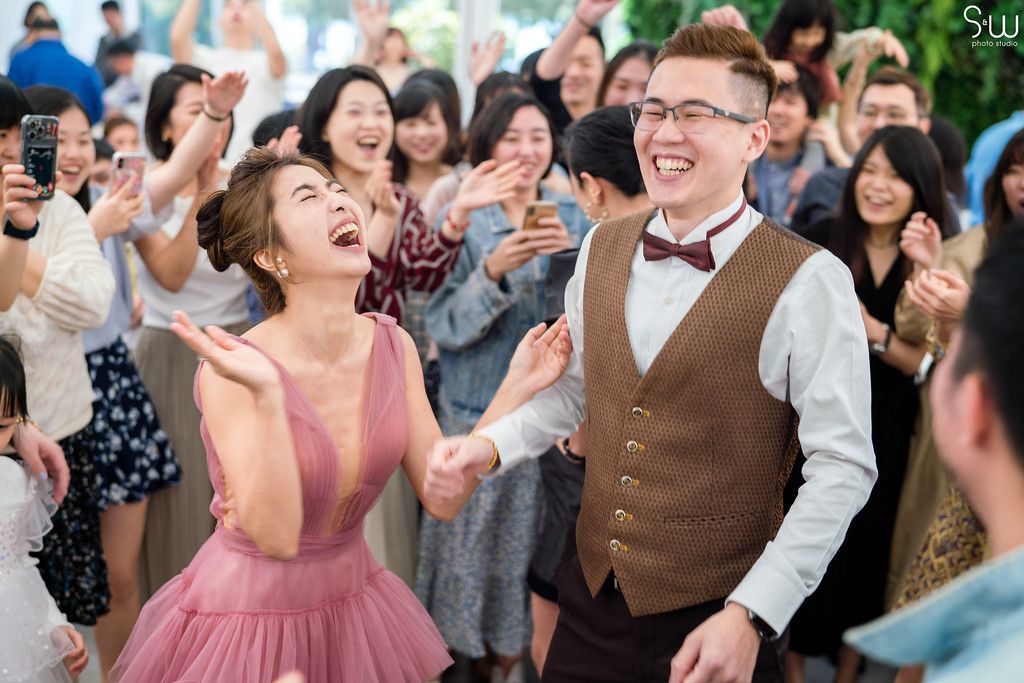 Guests celebrate enthusiastically at a reception under decorative greenery with a crowd cheering.