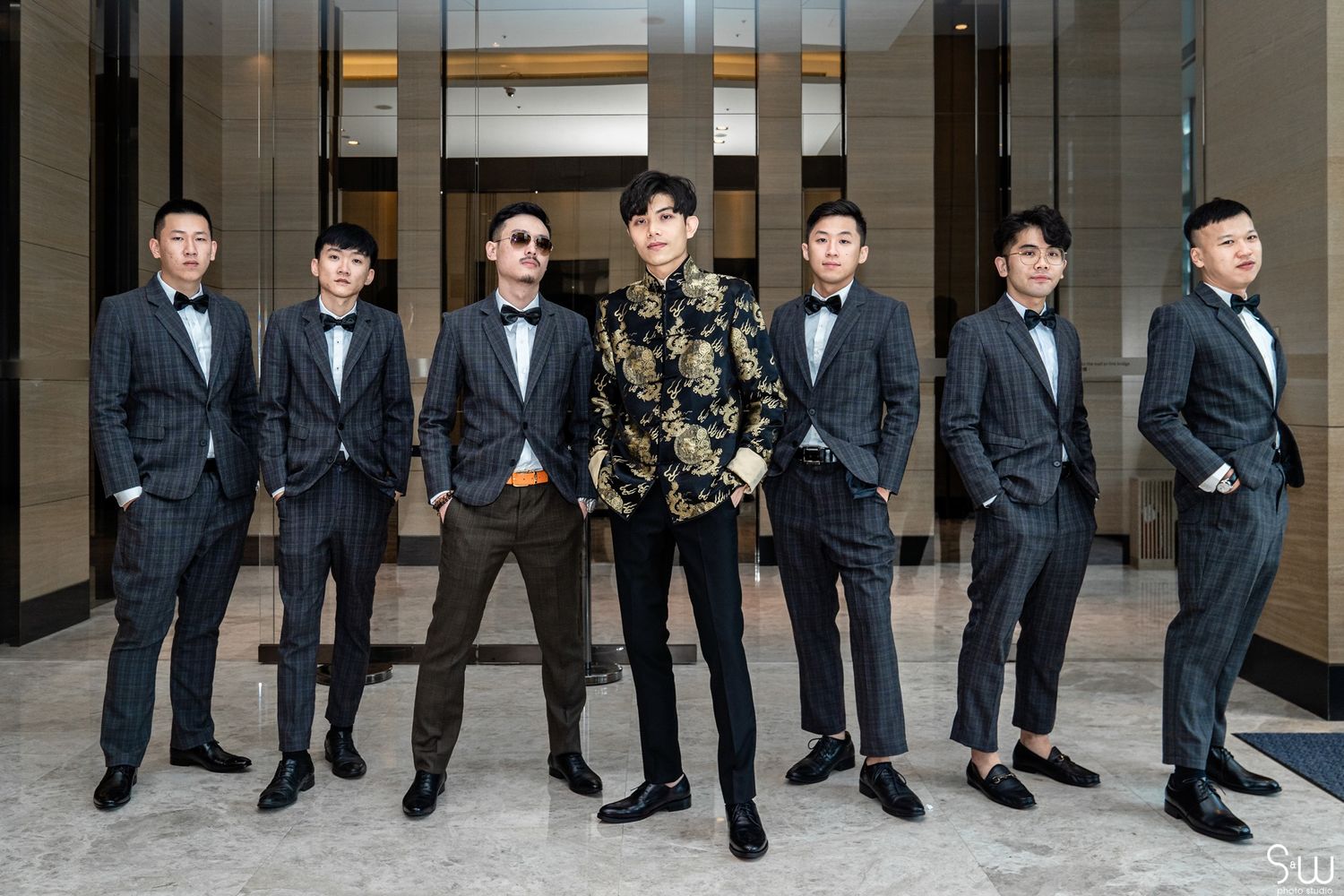 A group in formal attire poses elegantly in a modern lobby, with six wearing dark suits and one in a patterned shirt.