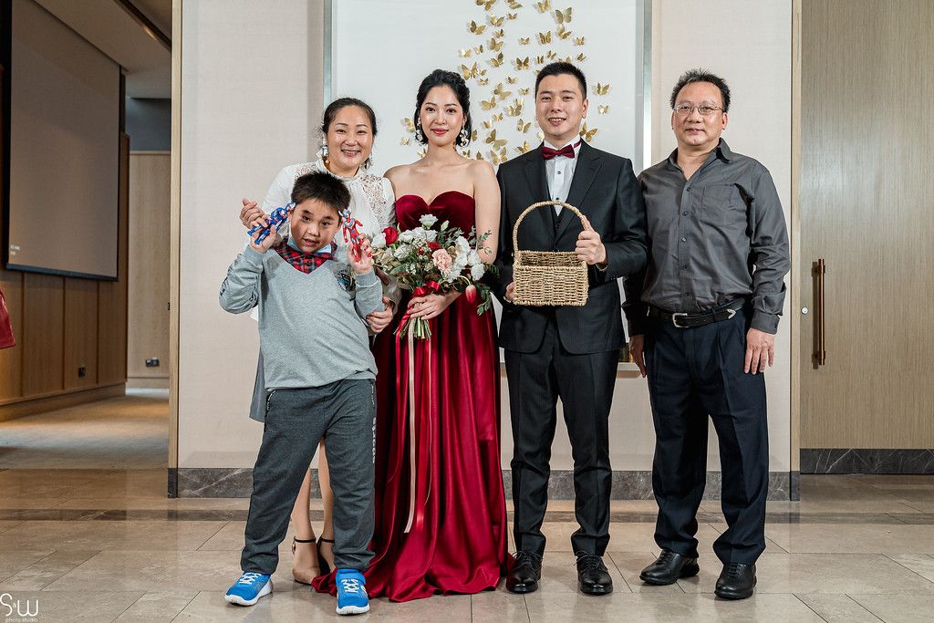 A wedding family portrait with a couple in formal attire standing with relatives in front of a decorative wall.