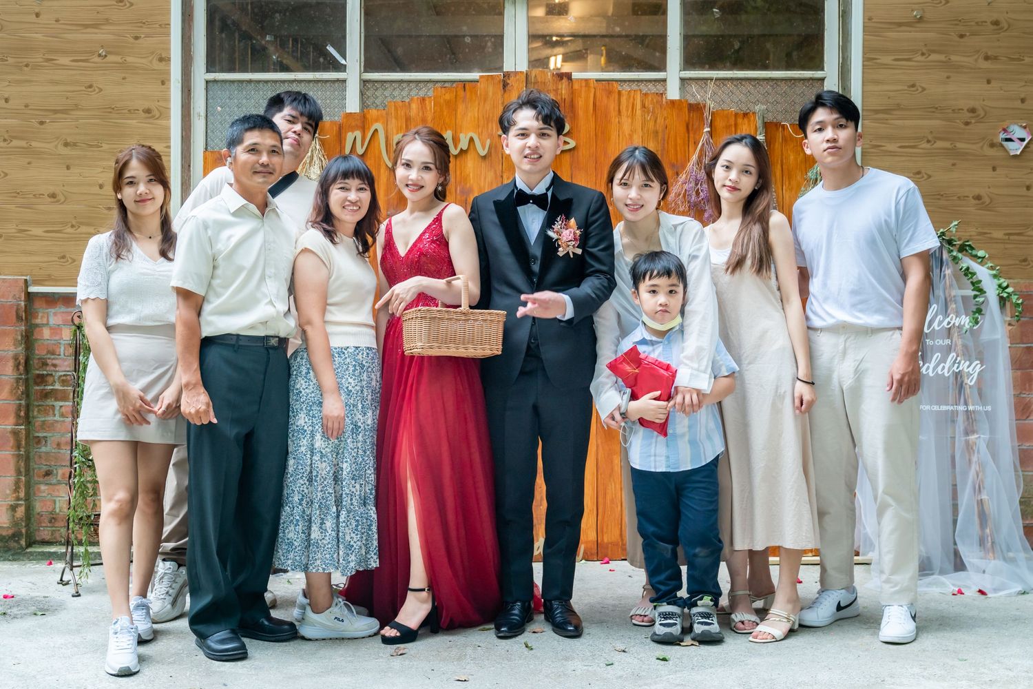 Large group in formal attire poses together against wooden wall with decorative elements and white dress display.