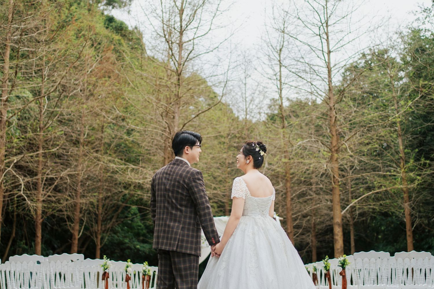 A couple in wedding attire stands together facing a forest backdrop with white chairs set up for an outdoor ceremony.