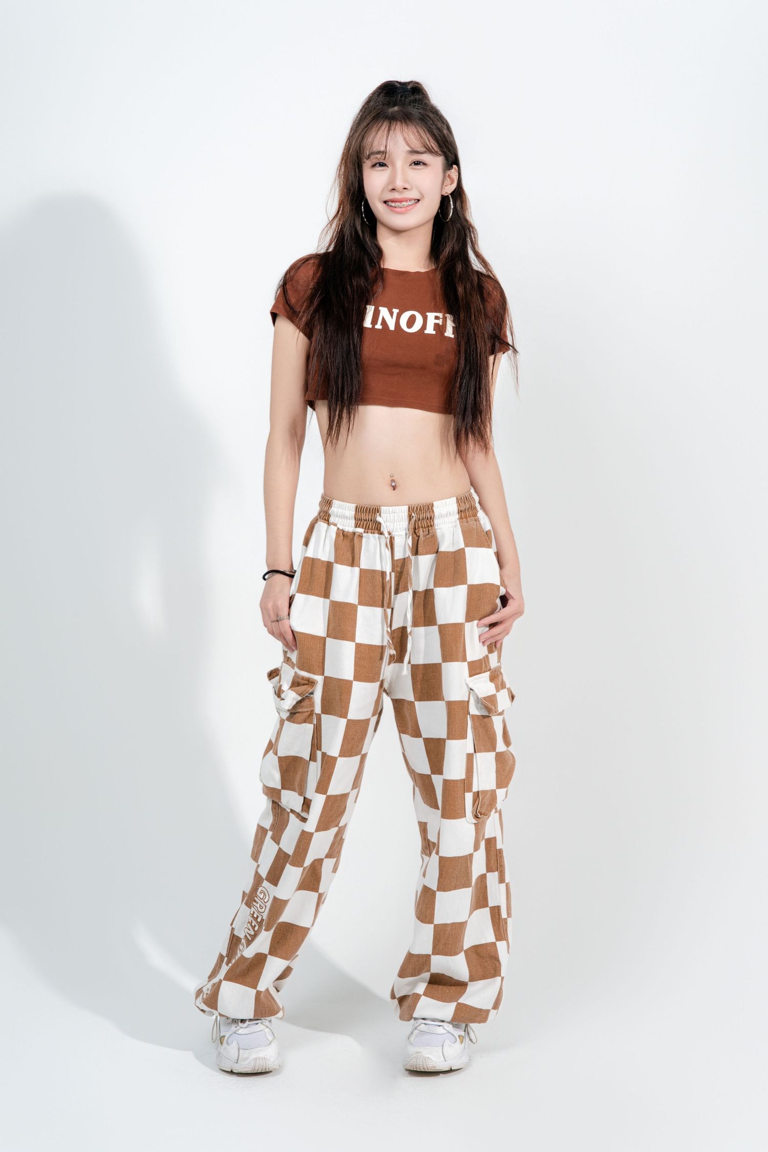 Model wearing brown crop top and checkered cargo pants in white studio setting against plain backdrop.