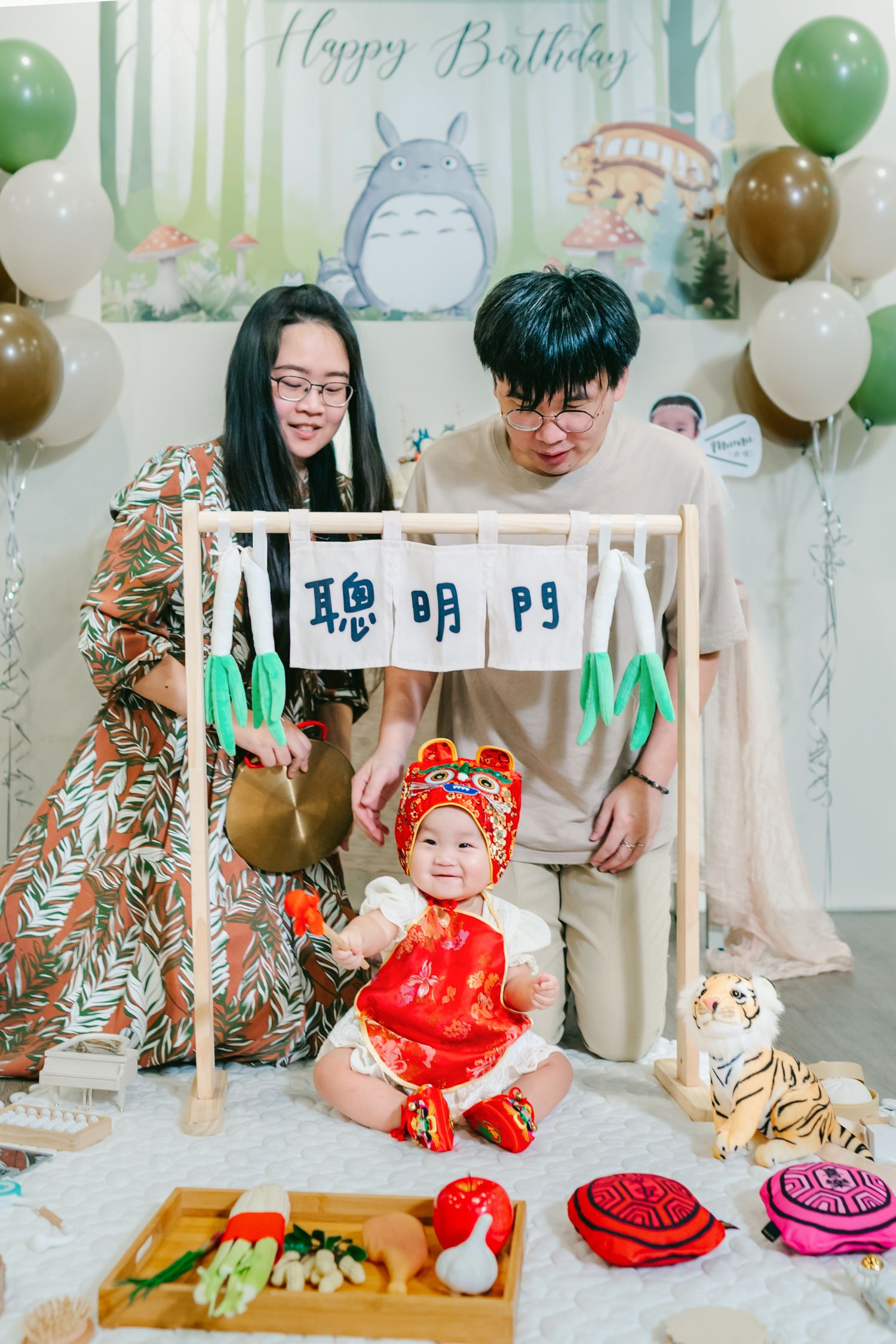 A baby celebrates a birthday in traditional red Chinese attire with Totoro-themed decorations and green and white balloons.