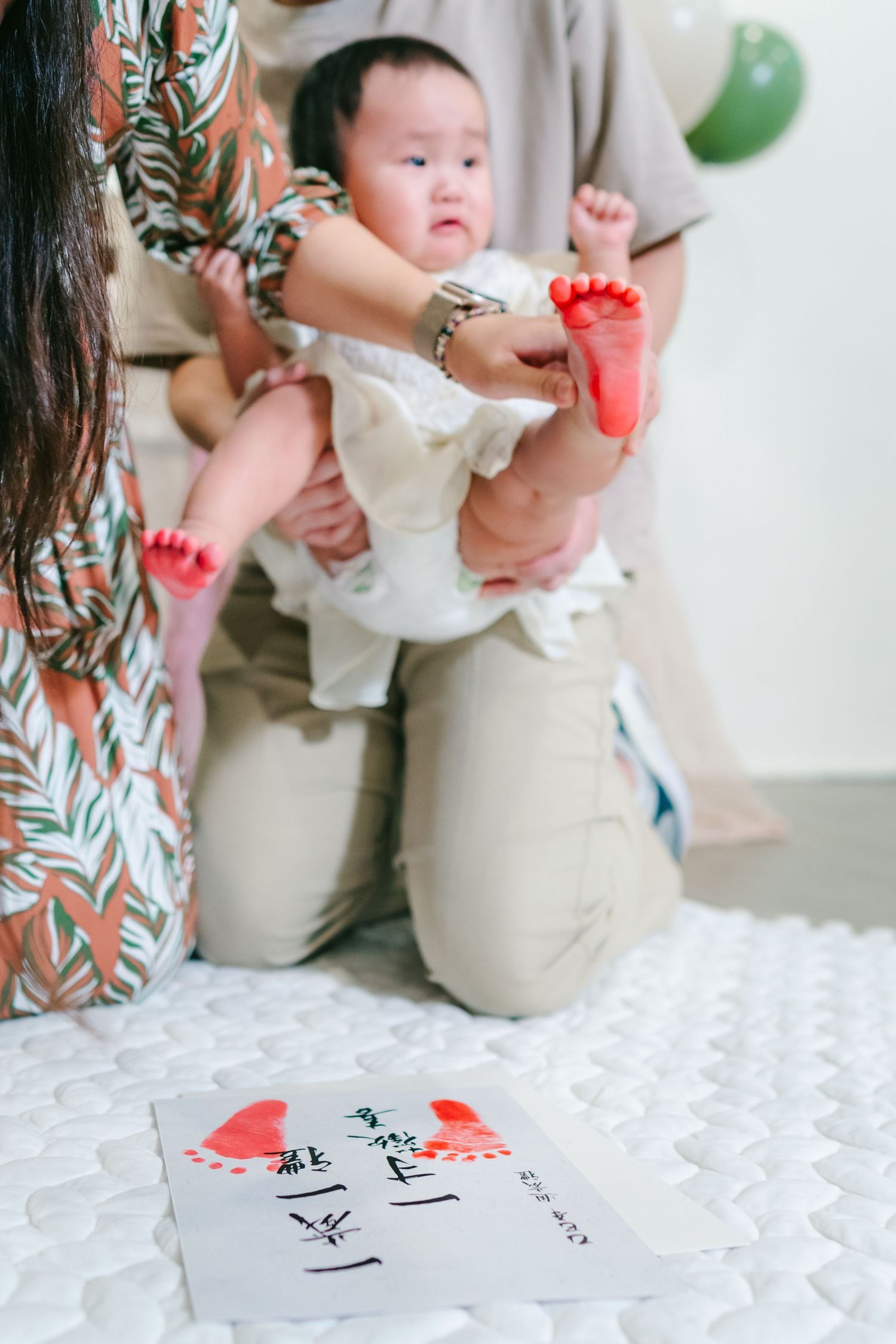 Baby in beige outfit sits on white textured surface with handwritten notes and red heart decorations visible nearby.