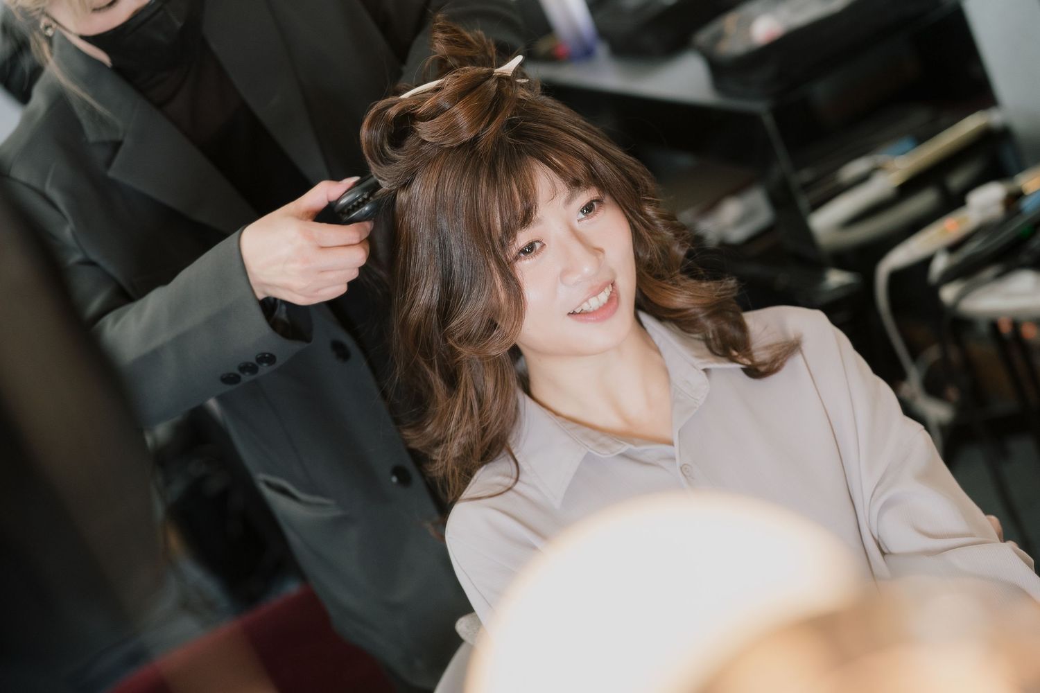Woman smiling while getting her hair styled at a salon, sitting in a chair with a stylist working behind her.