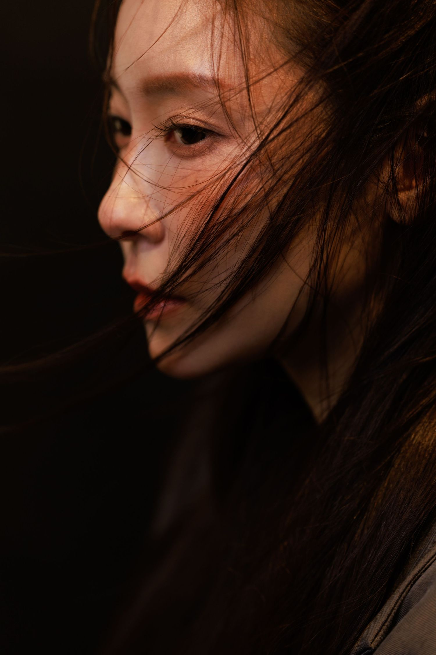 A young Asian woman in profile with dark hair blowing across her face against a dark background.