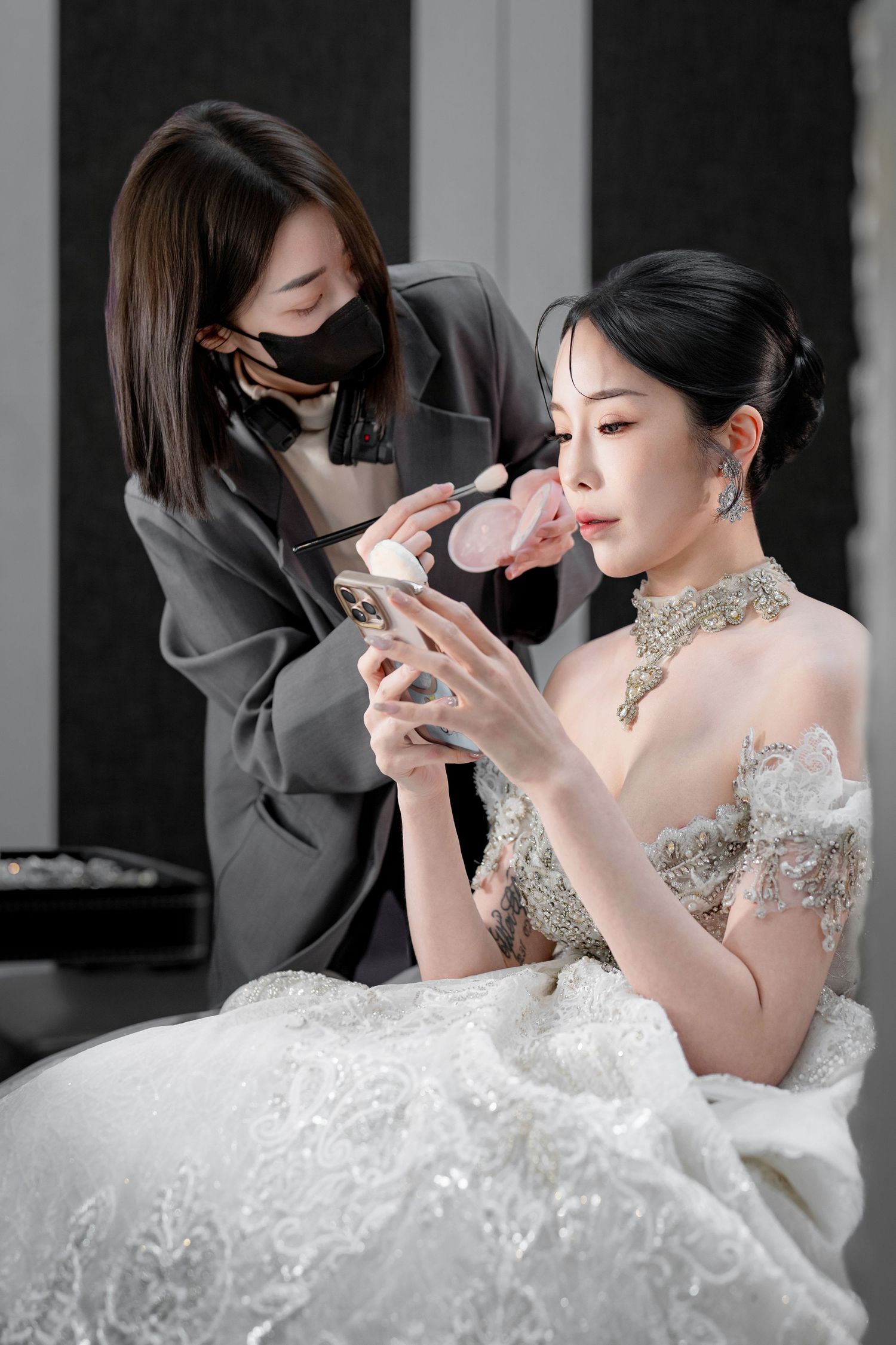 Makeup artist applies cosmetics to bride wearing elegant white lace gown and crystal necklace during wedding preparation.