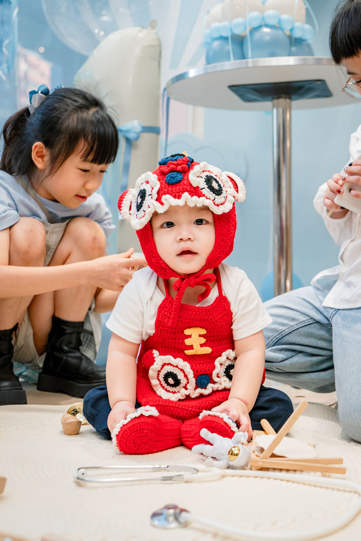 A small child in a red knitted outfit plays at a table with toys while others look on in a bright indoor setting.