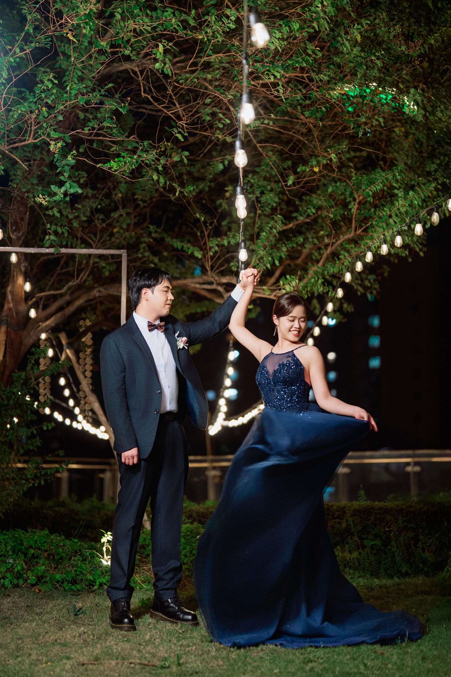 A couple in formal attire dances playfully under string lights in a garden setting at dusk.