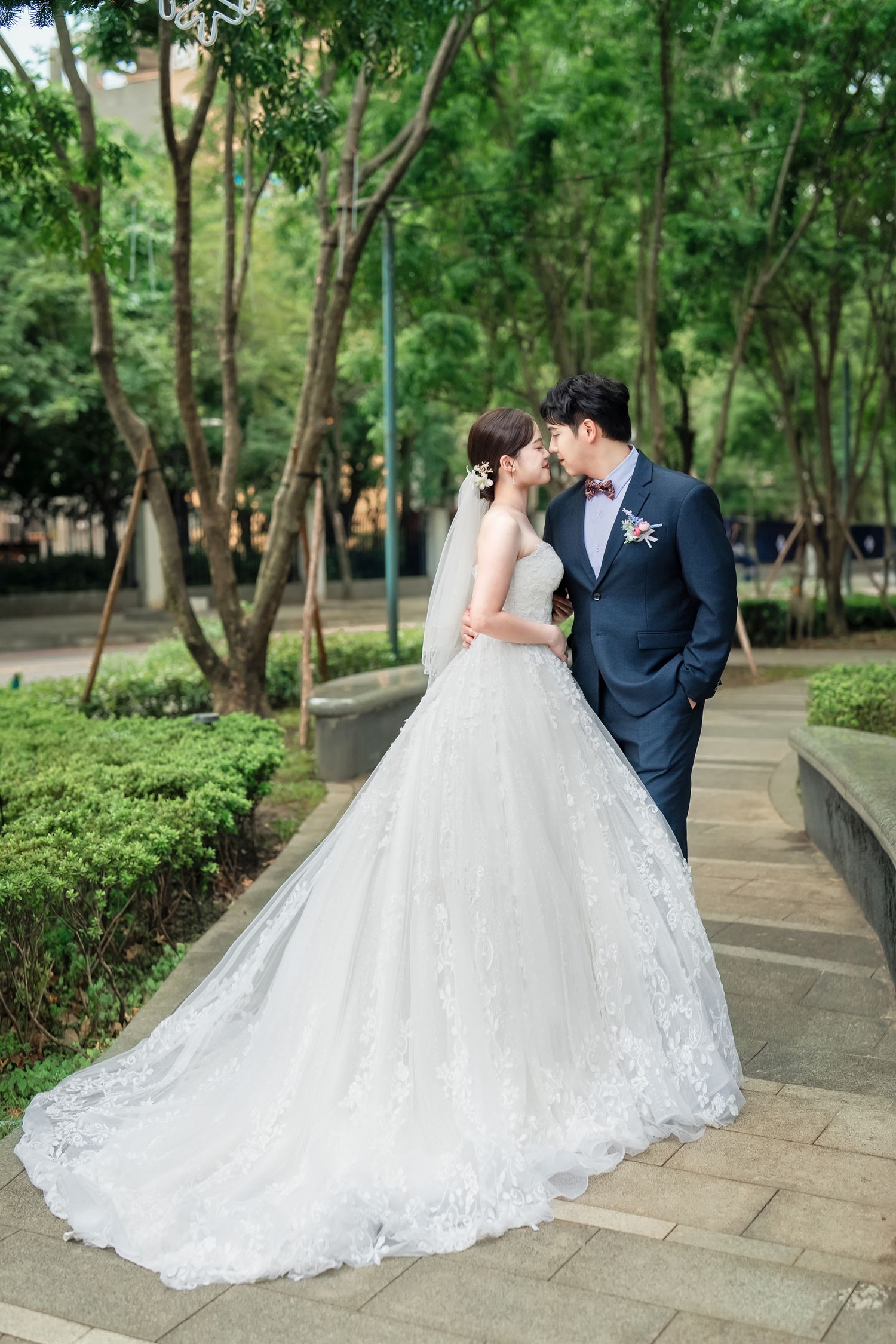 A bride in a flowing lace wedding gown with long train stands with groom in navy suit on garden path surrounded by trees.