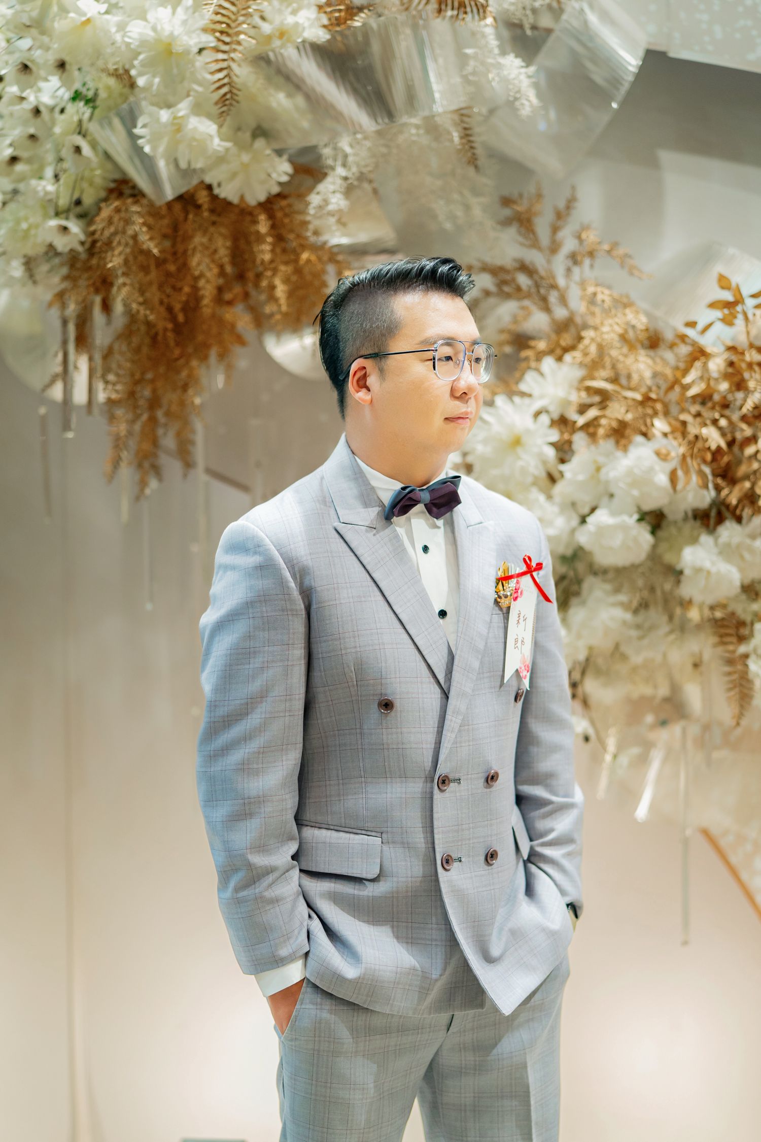 A person in a light gray suit stands in front of a wedding backdrop decorated with white flowers and dried foliage.