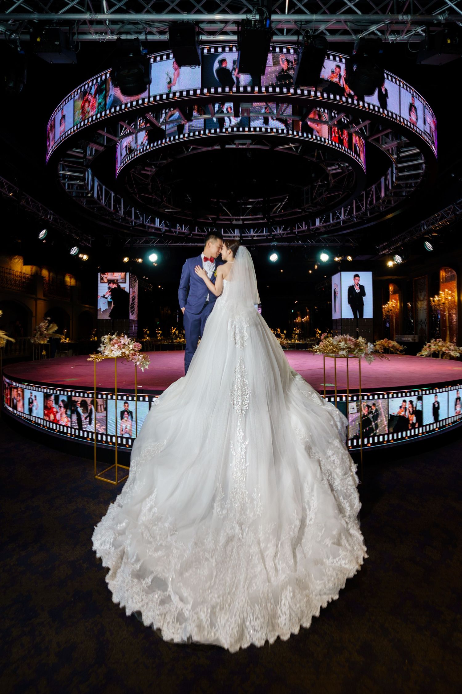Elegant wedding scene in dark venue with circular film strip displays and dramatic long lace train on white bridal gown.