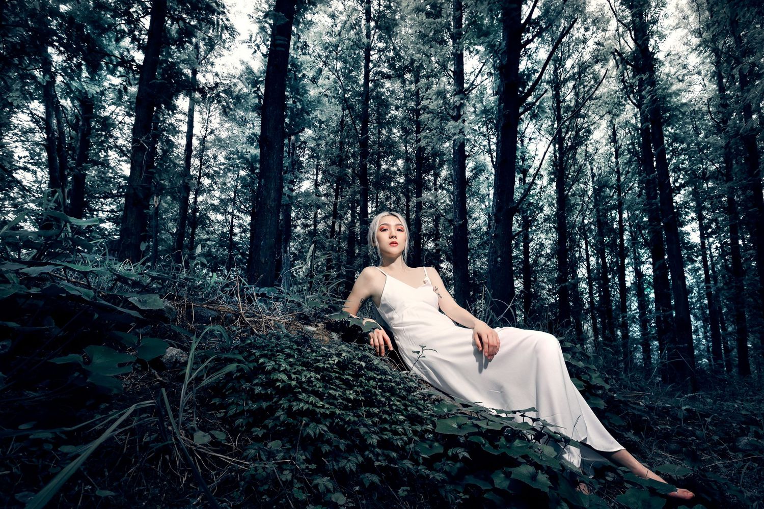 A figure in a flowing white dress reclines gracefully against a mossy slope in a dark, mysterious forest setting.